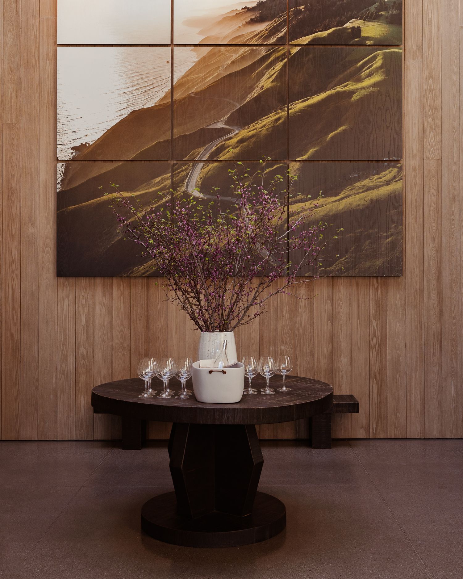 A decorative vase with purple flowers sits on a dark wooden table beneath a coastal landscape artwork on a wood-paneled wall.