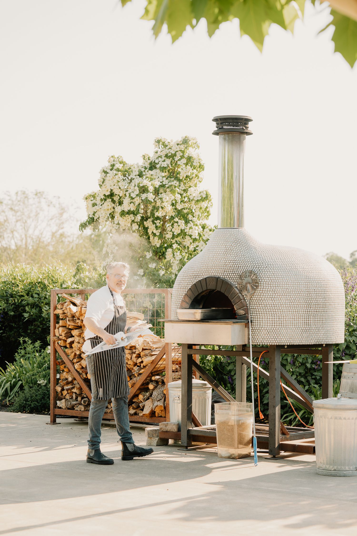 A wood-fired pizza oven with tiled mosaic exterior sits on a patio next to stacked firewood.