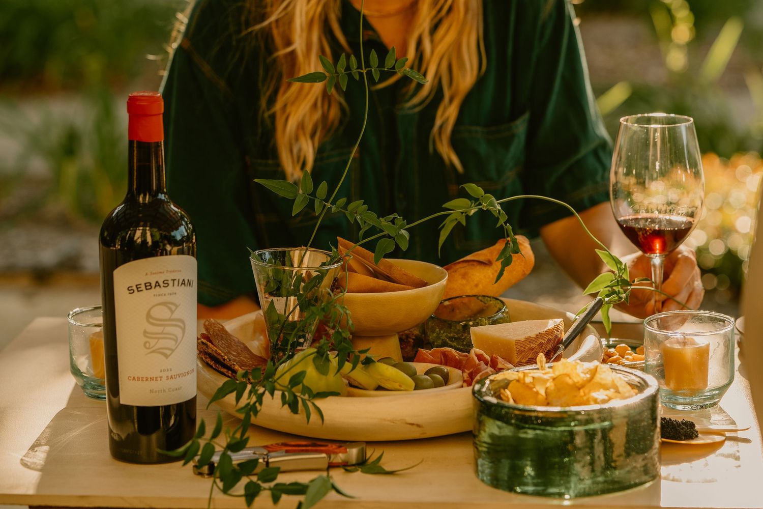 Woman enjoying a charcuterie spread outdoors with Cedartan red wine bottle and glass on a wooden table.