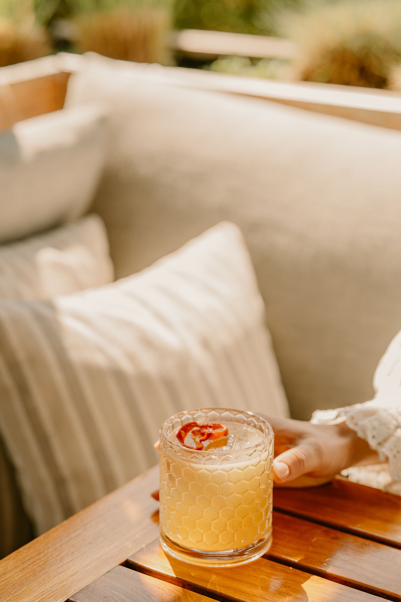 A golden cocktail garnished with dried flowers sits on a wooden table beside a cozy outdoor sofa with cream pillows.
