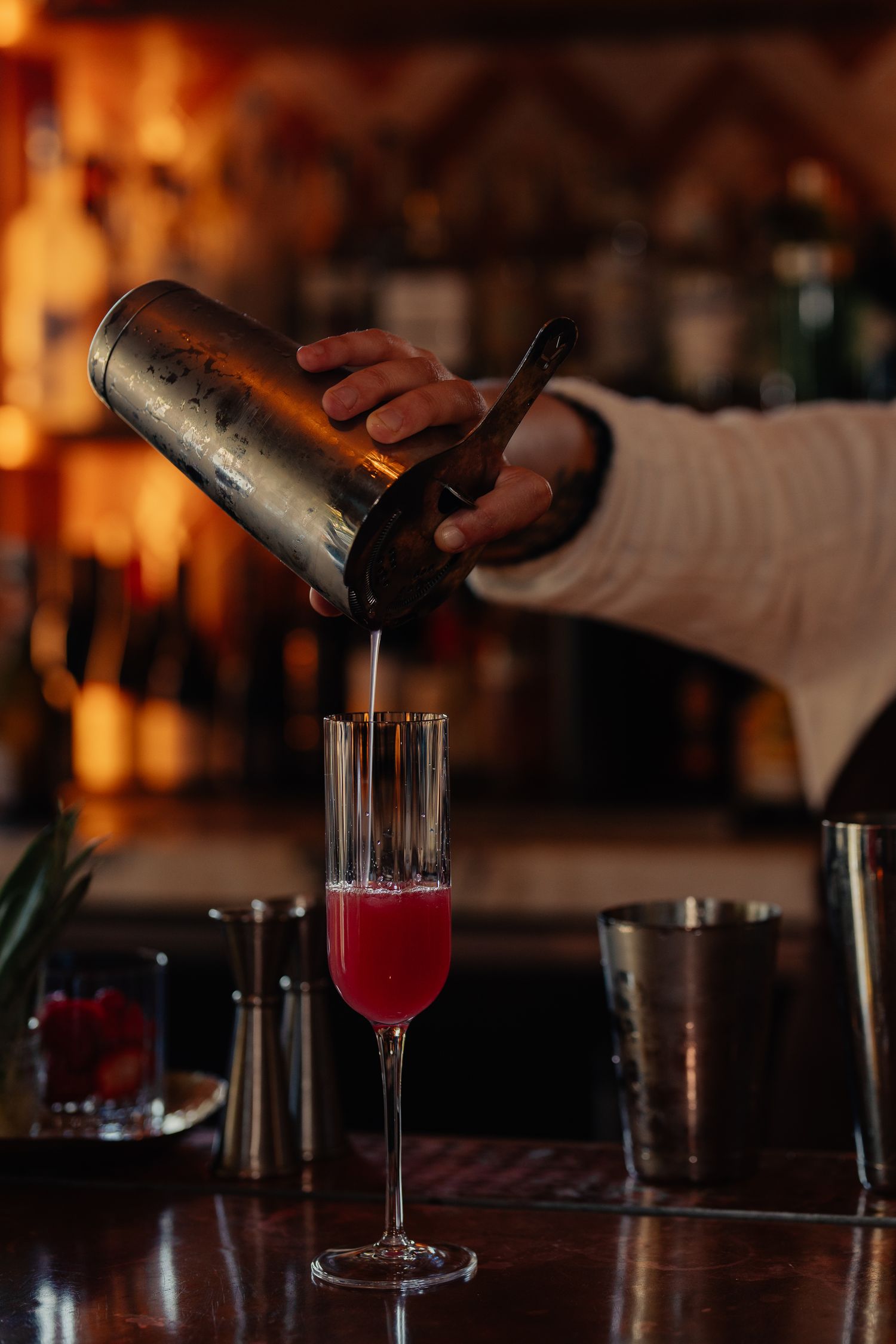 Bartender pouring a red cocktail from a shaker into a champagne flute in a dimly lit bar setting.