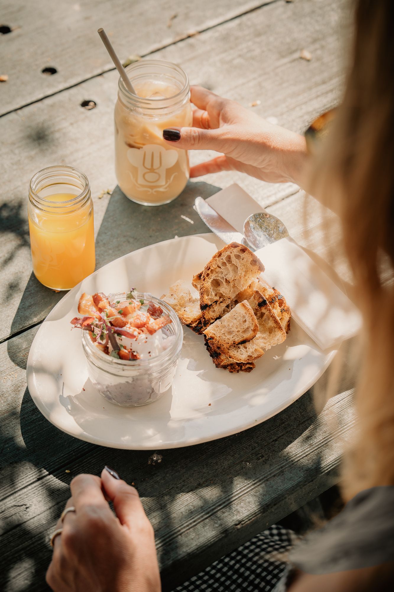 Woman enjoying outdoor brunch with toasted bread, fresh salad, orange juice, and iced coffee on a wooden table.