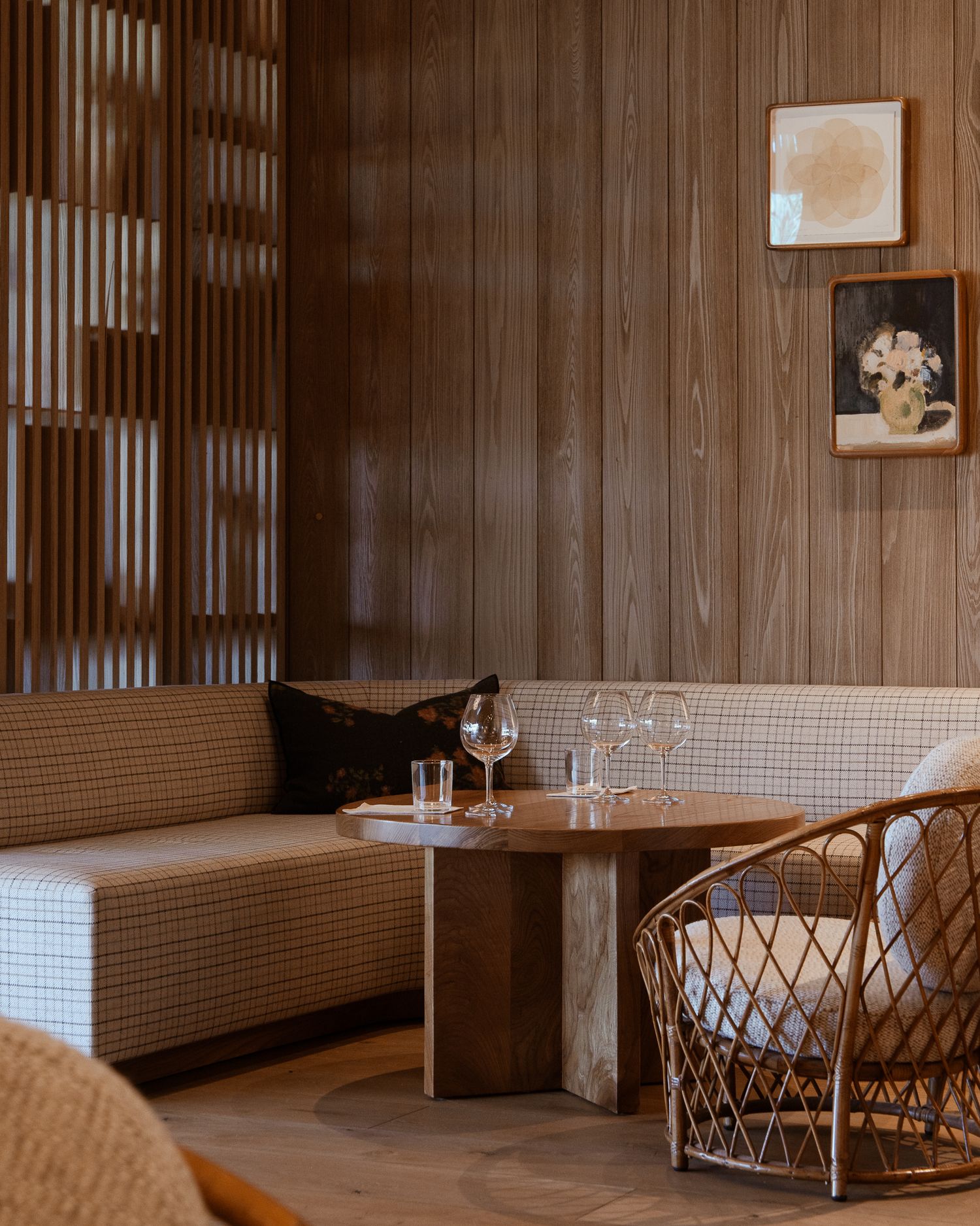 Modern restaurant interior with wooden walls, built-in bench seating, rattan chair and wine glasses on a round table.