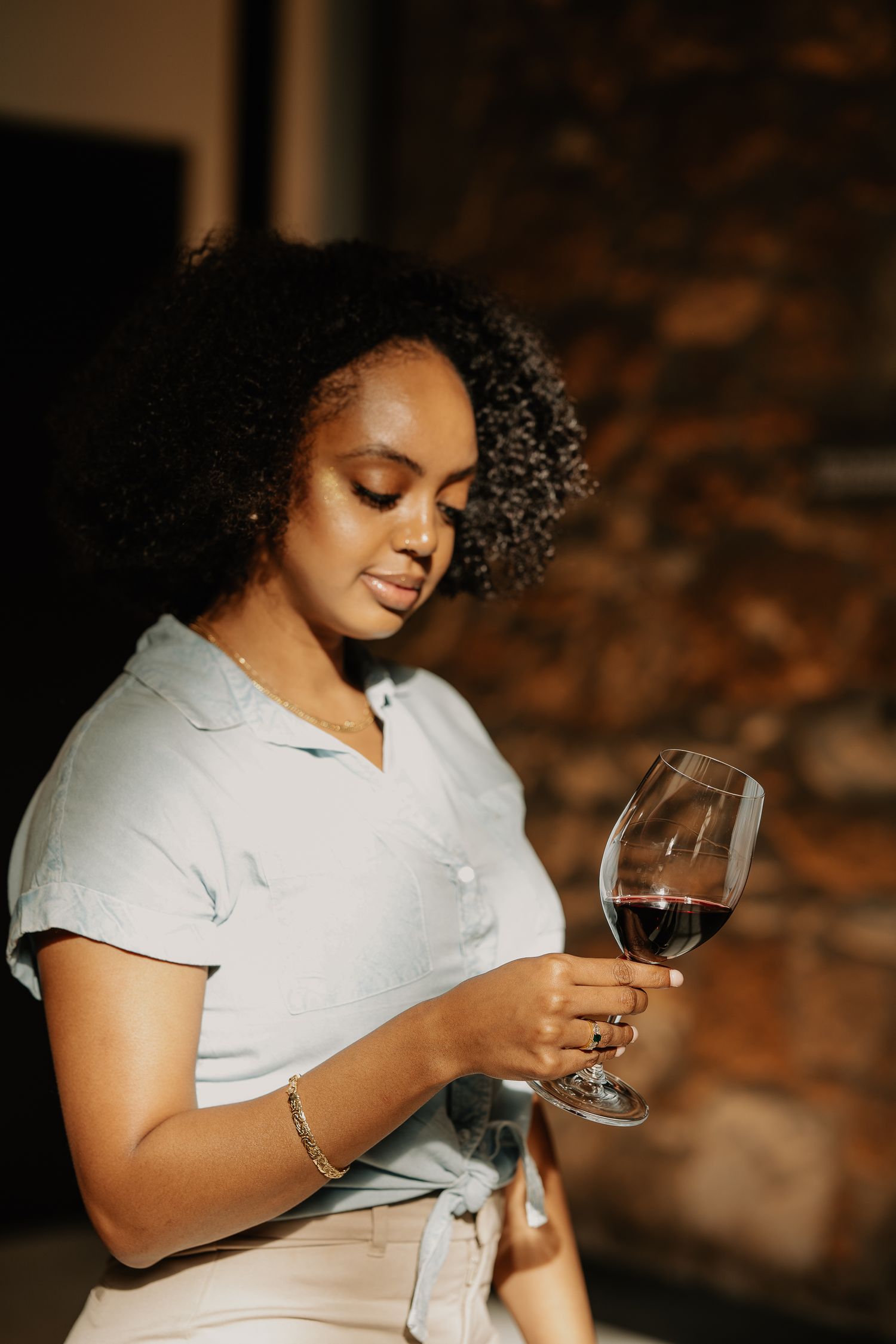 Young Black woman with curly hair examines a glass of red wine in warm, dimly lit setting.