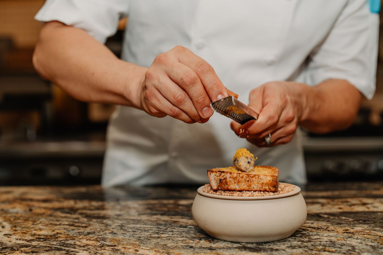 Chef seasoning a dish in a white bowl on a rustic wooden surface with fresh herbs.