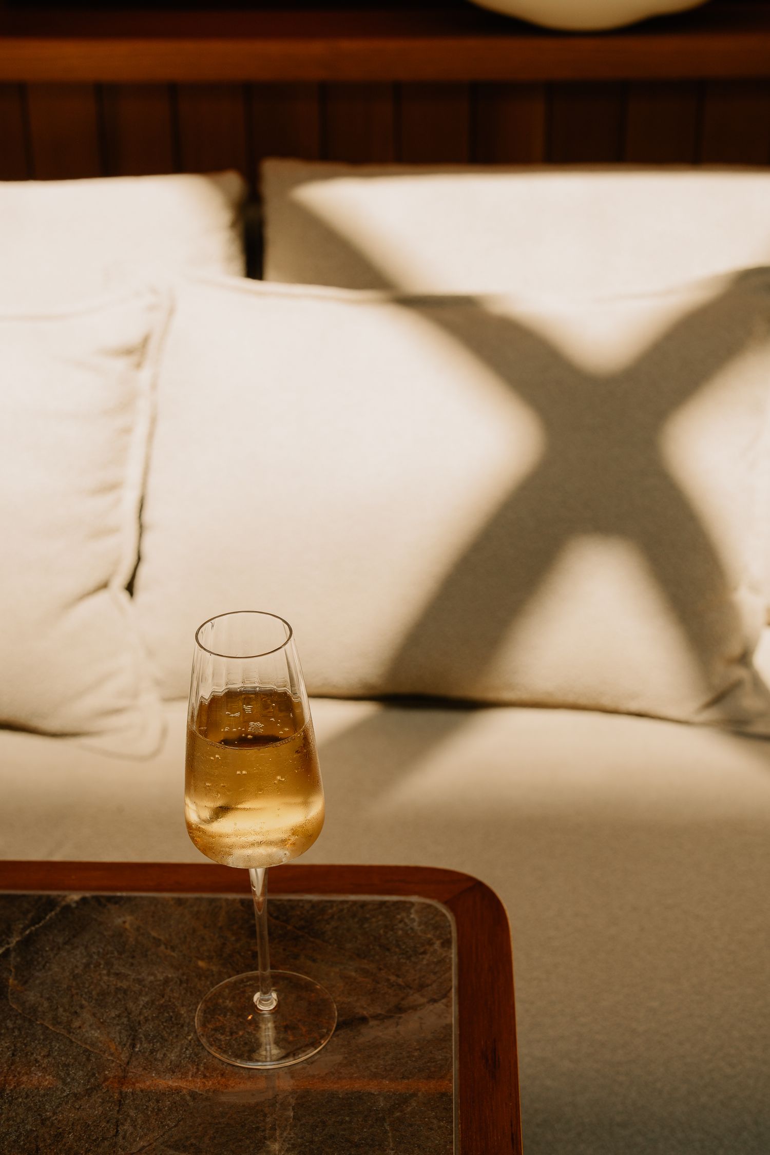 A glass of white wine sits on a wooden table against white pillows with dramatic window shadow patterns.