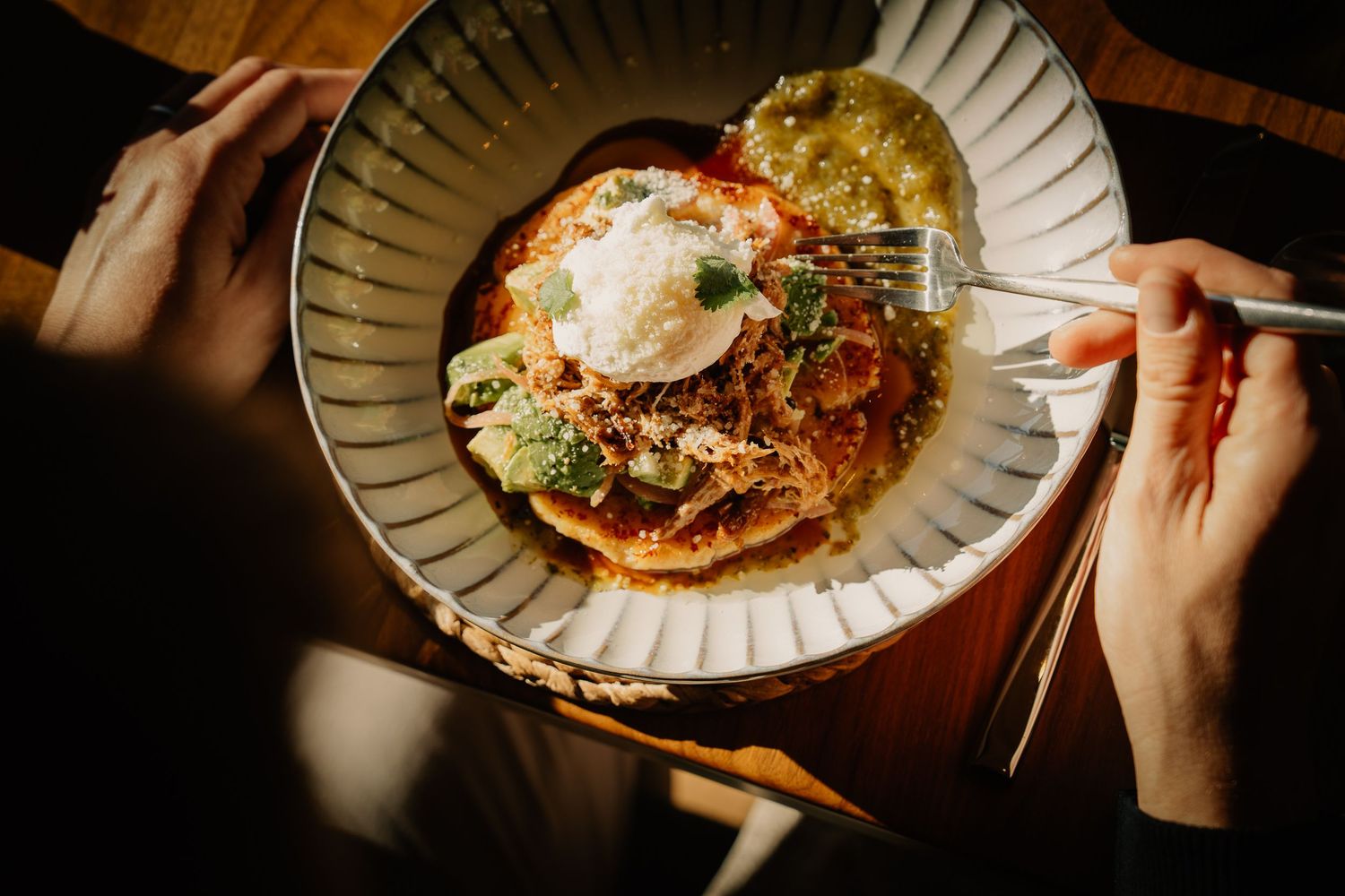 A hand holds chopsticks over a steaming bowl of ramen noodle soup garnished with fresh herbs and toppings.