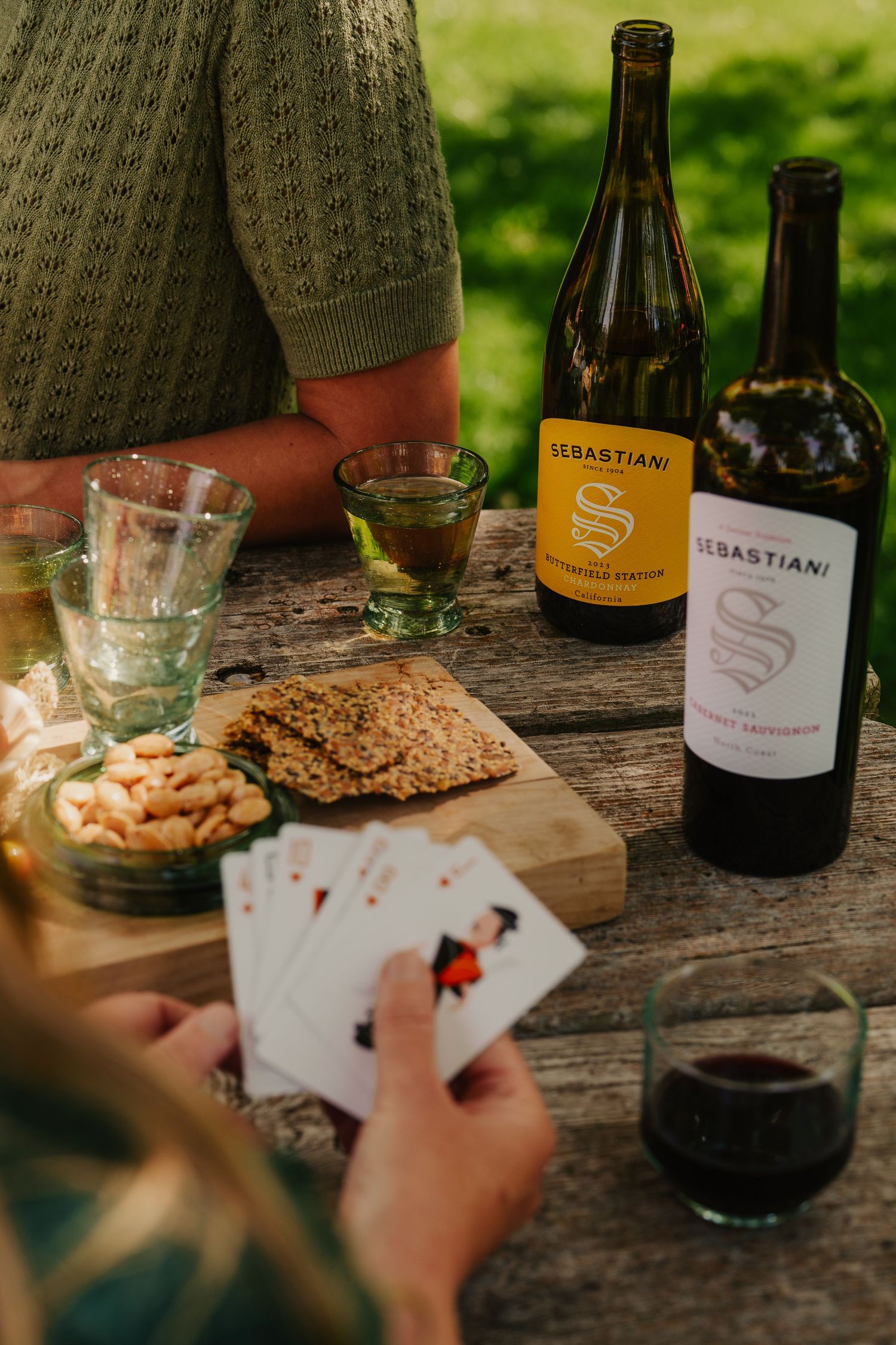 Friends playing cards with Sebastiani Chardonnay and Cabernet Sauvignon bottles at rustic outdoor table.