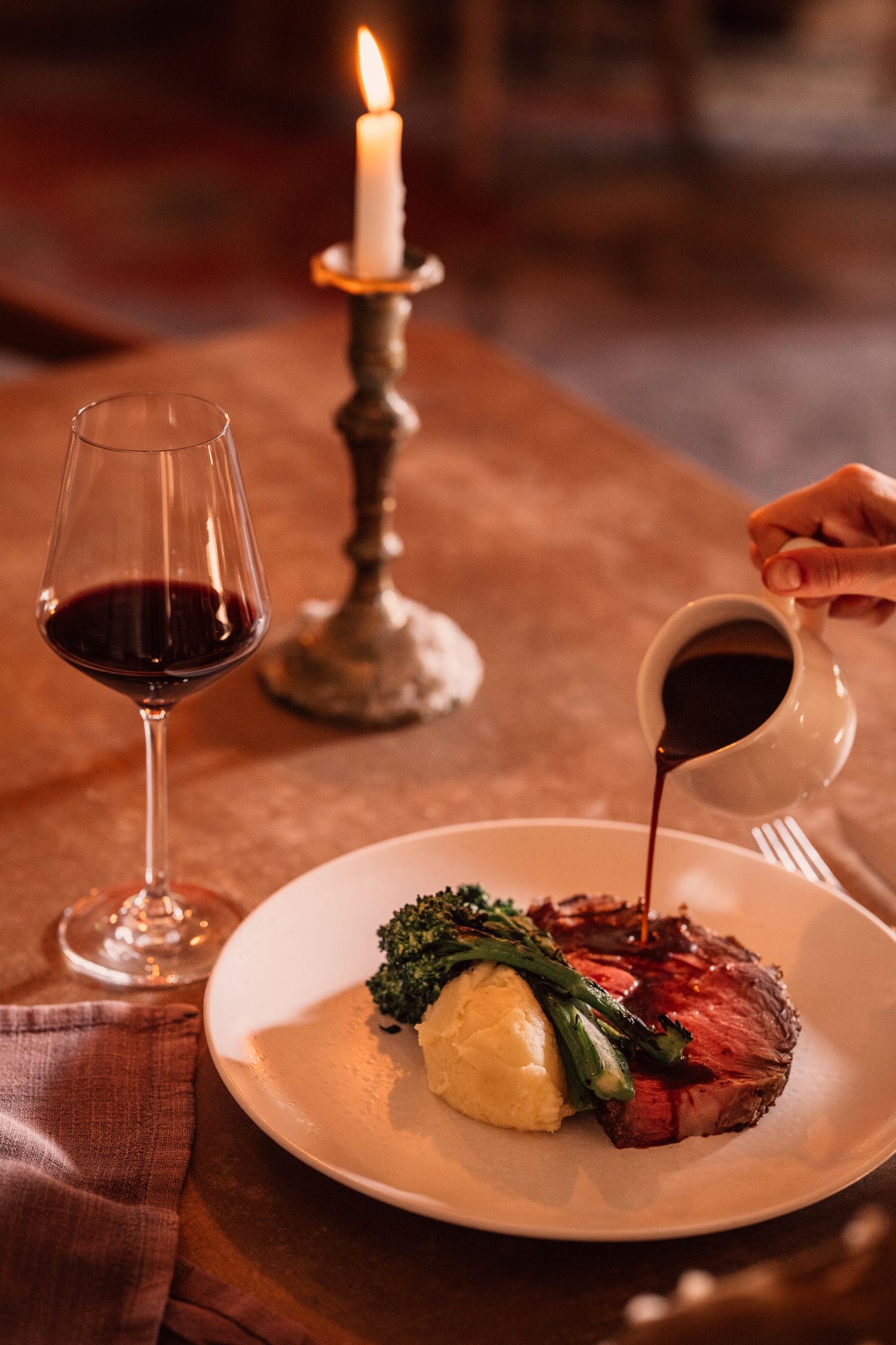 Fine dining plate with meat and vegetables being drizzled with sauce, served alongside red wine and candlelight.