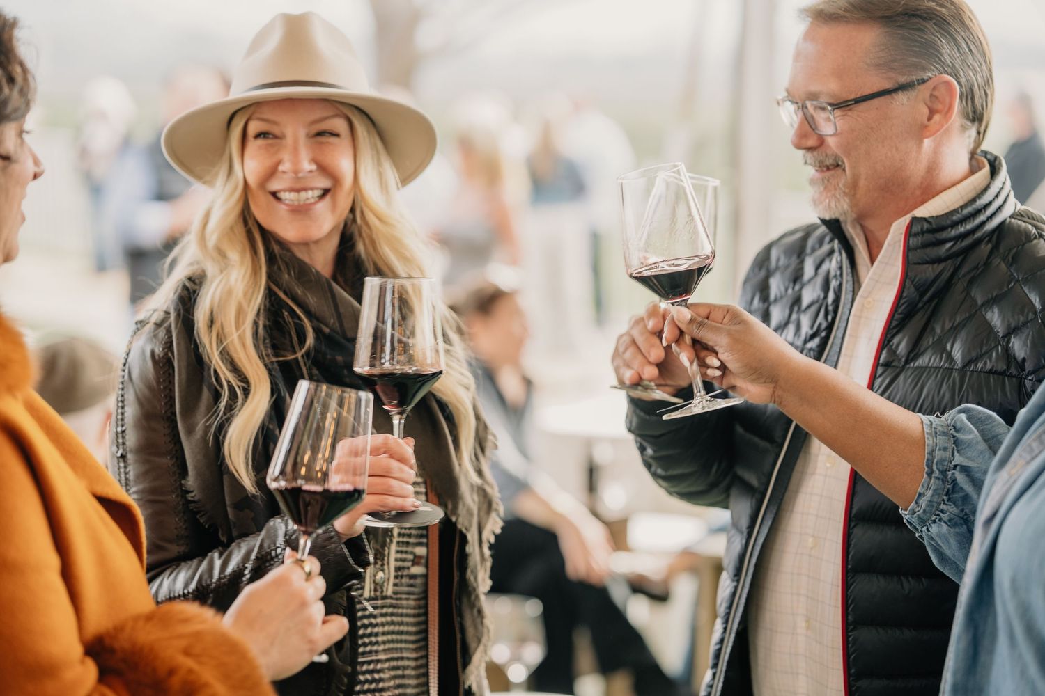 Group of smiling guests enjoying red wine at an outdoor wine tasting event under a tent.