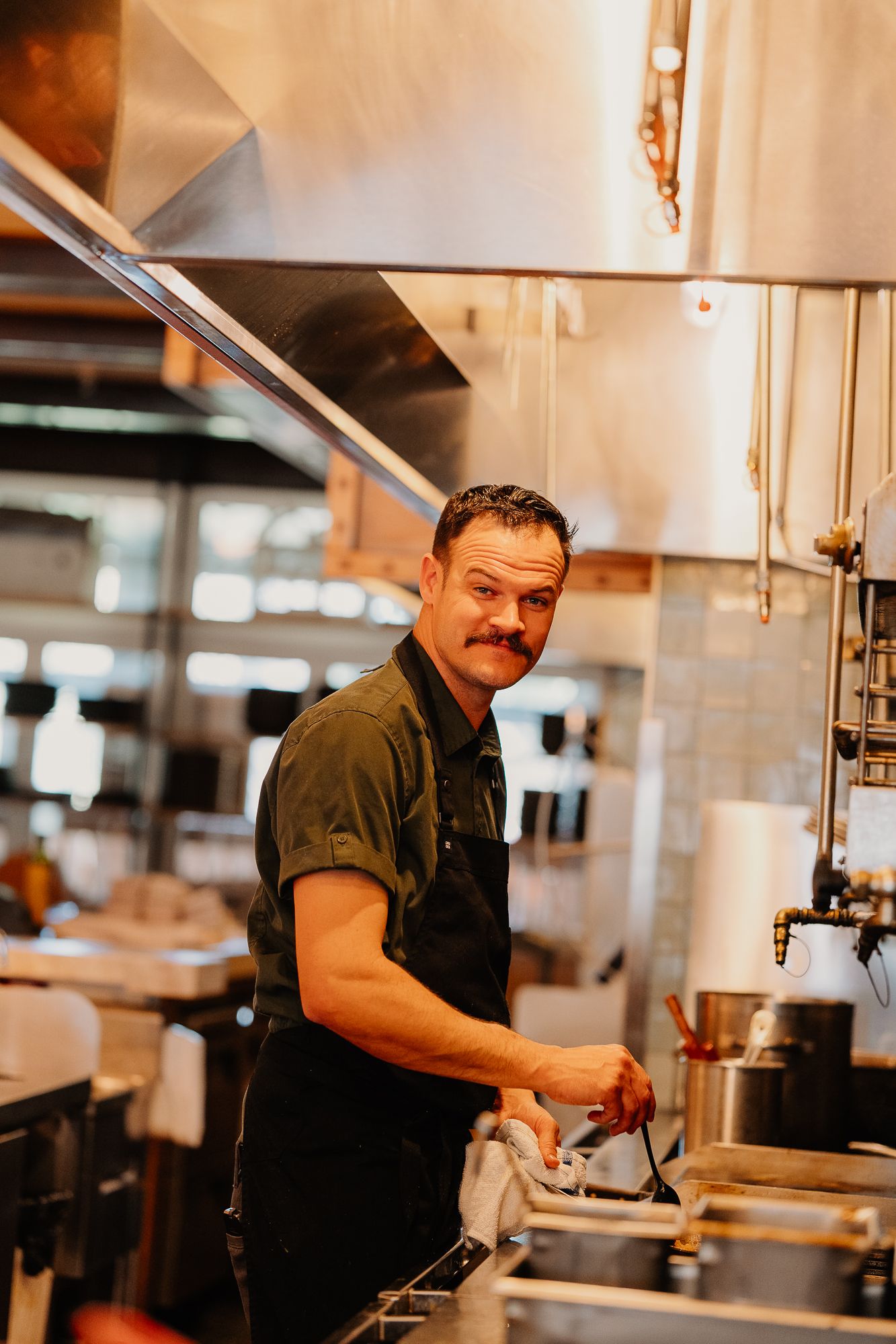 Chef working in a professional kitchen wearing a black apron, preparing food at the stove.