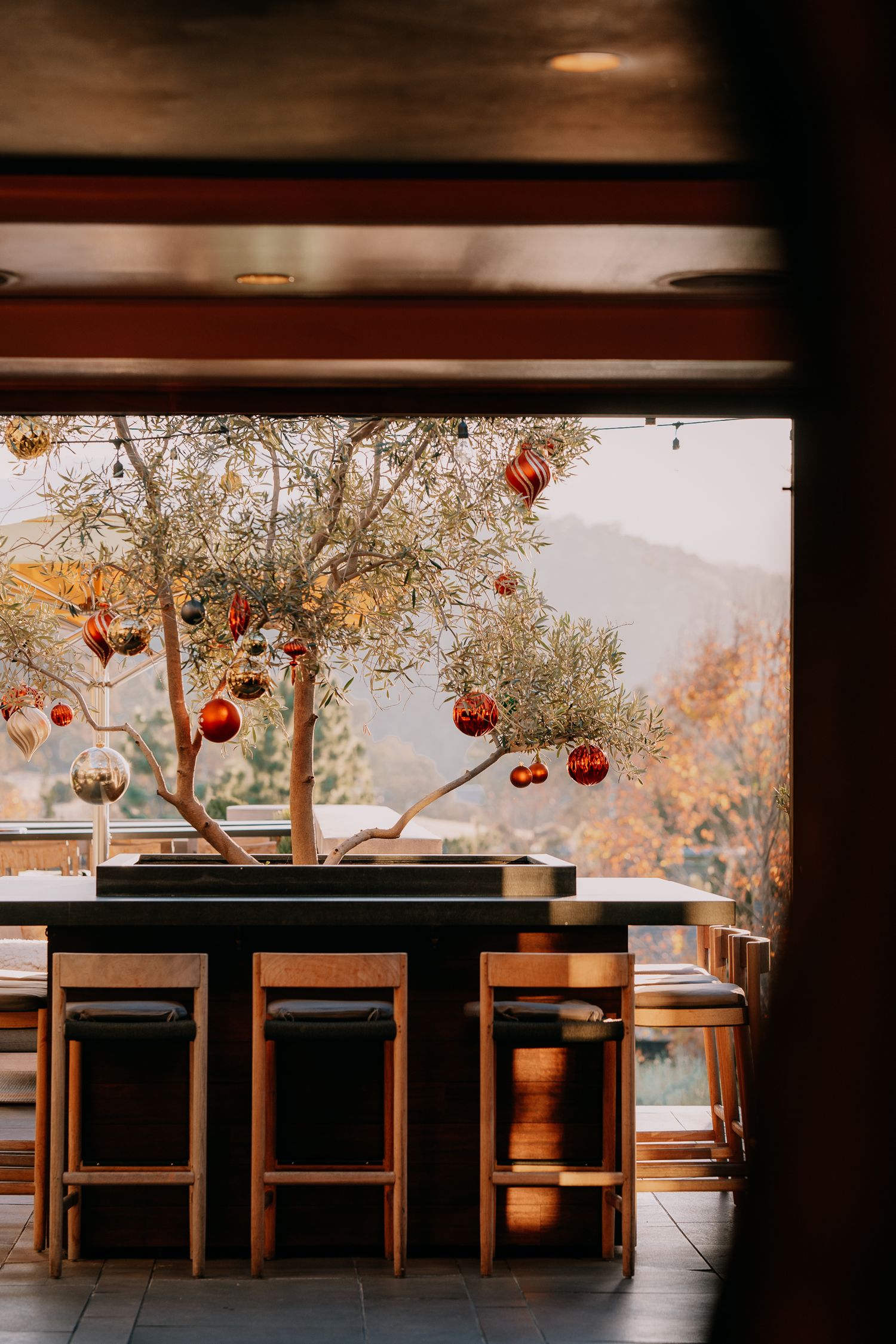 Red Christmas ornaments hang from bare tree branches above a wooden bar counter at an outdoor restaurant terrace.