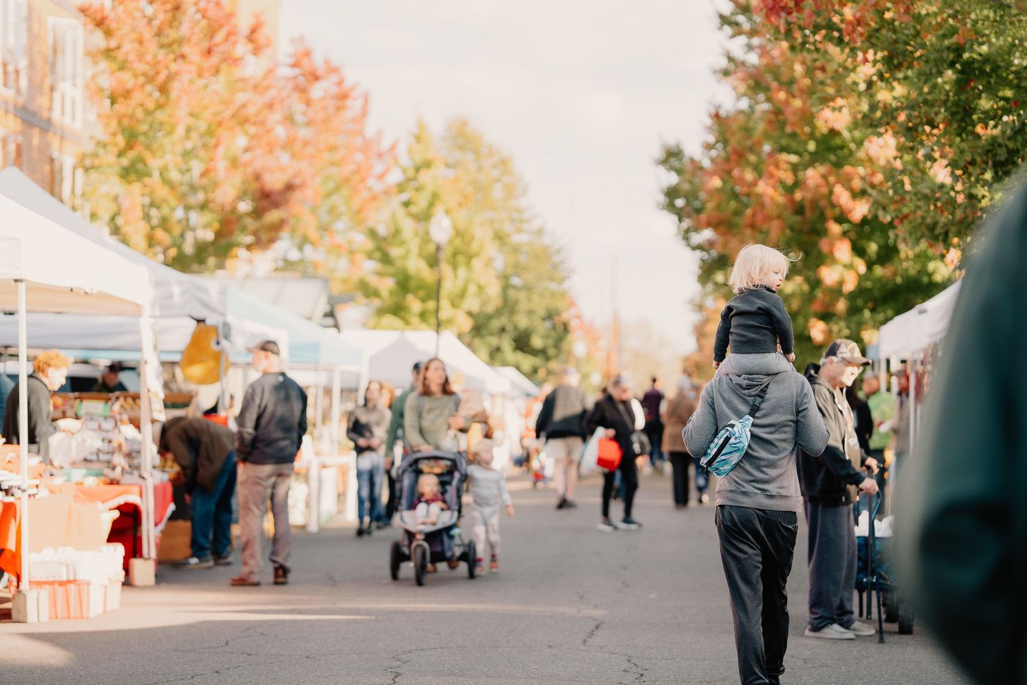 People walk through a lively outdoor market with white tents and fall foliage on a sunny autumn day.