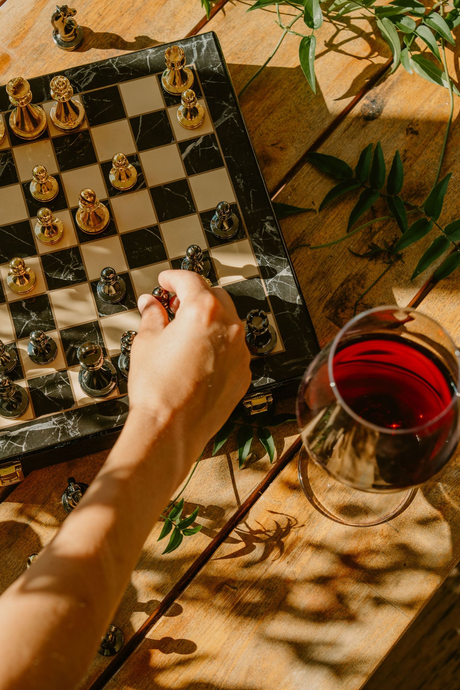 Hand moving a chess piece on a marble board beside a glass of red wine on a sunlit wooden table.