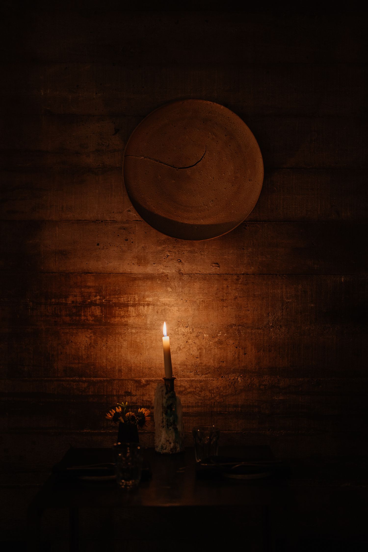 A single lit candle glows warmly against a rustic wooden background with a round shape casting shadows above.