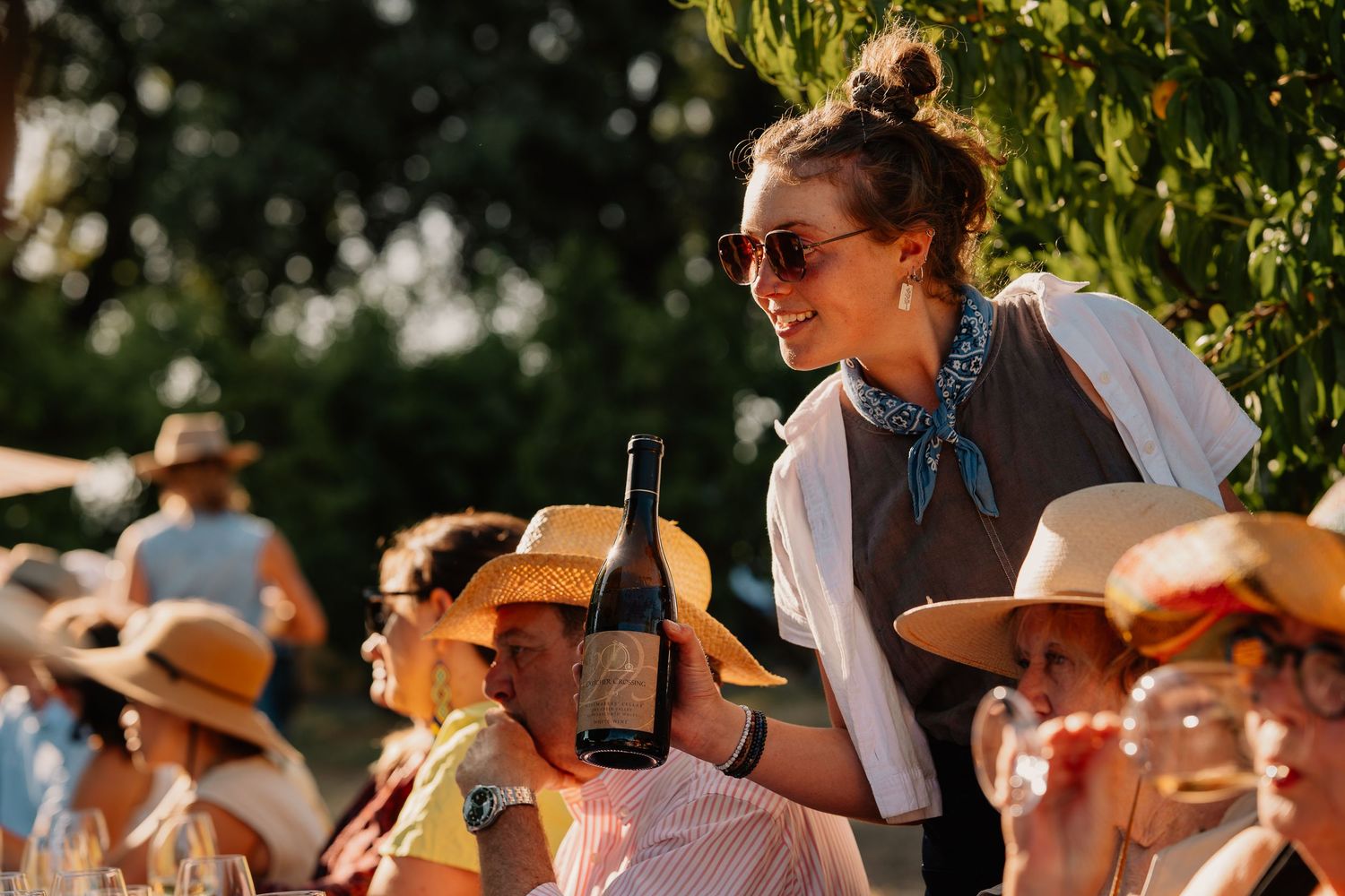 People enjoying an outdoor wine tasting event in the late afternoon sun wearing hats and sunglasses.