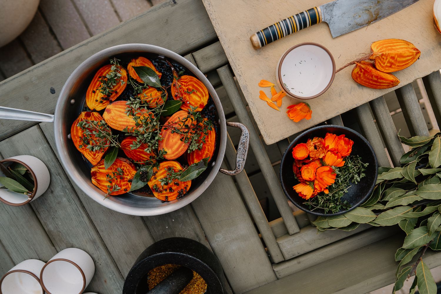 Overhead view of roasted tomatoes and herbs in a silver pan on a rustic wooden table with a knife and bowl.