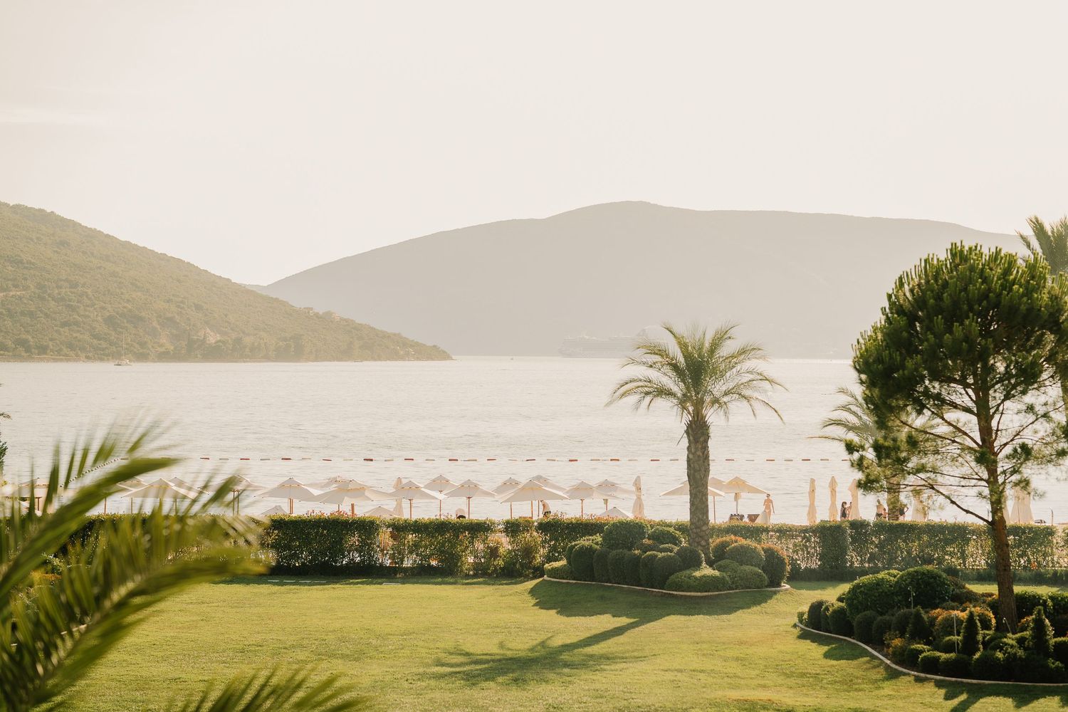 Lush green lawn with palm trees overlooking a serene bay, beach umbrellas, and mountain backdrop at a luxury resort.
