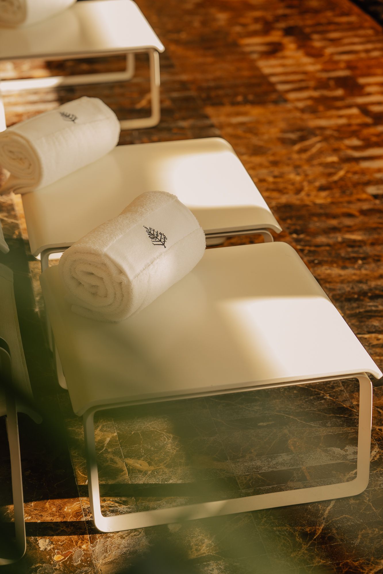 White rolled towels with embroidered logo on modern lounge chairs in warm sunlight at a luxury spa.