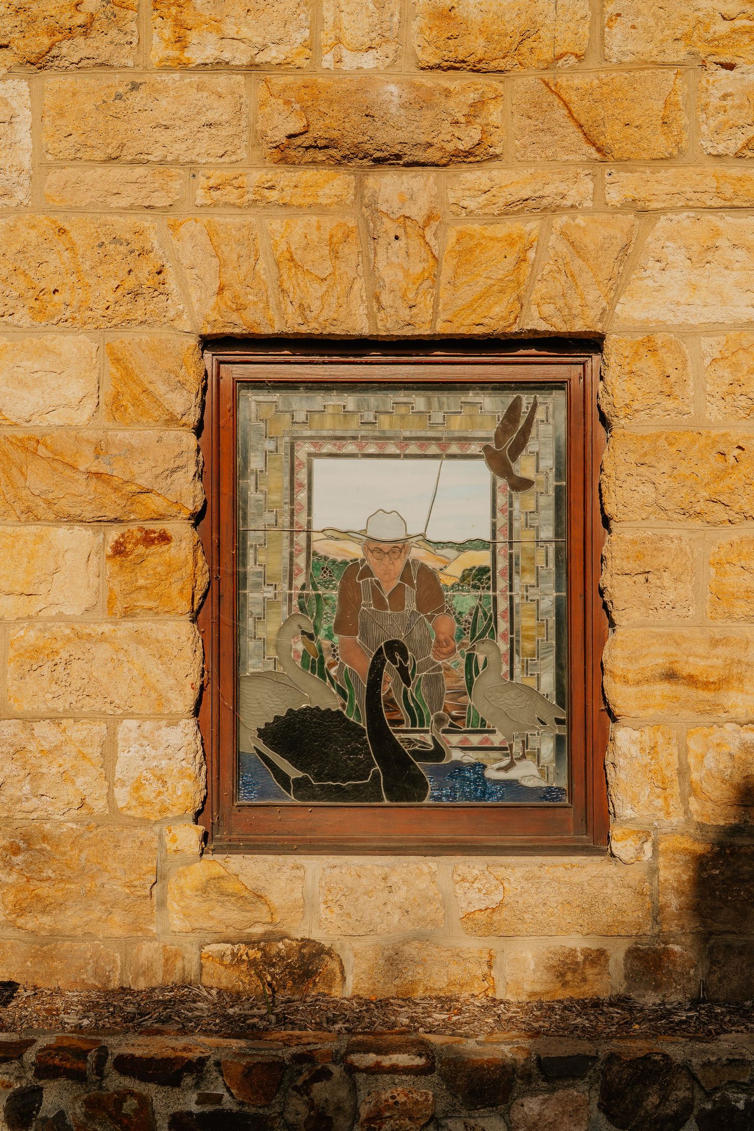 Mosaic artwork in wooden frame set into a sandstone wall depicting a farmer with black swans in a rural landscape.