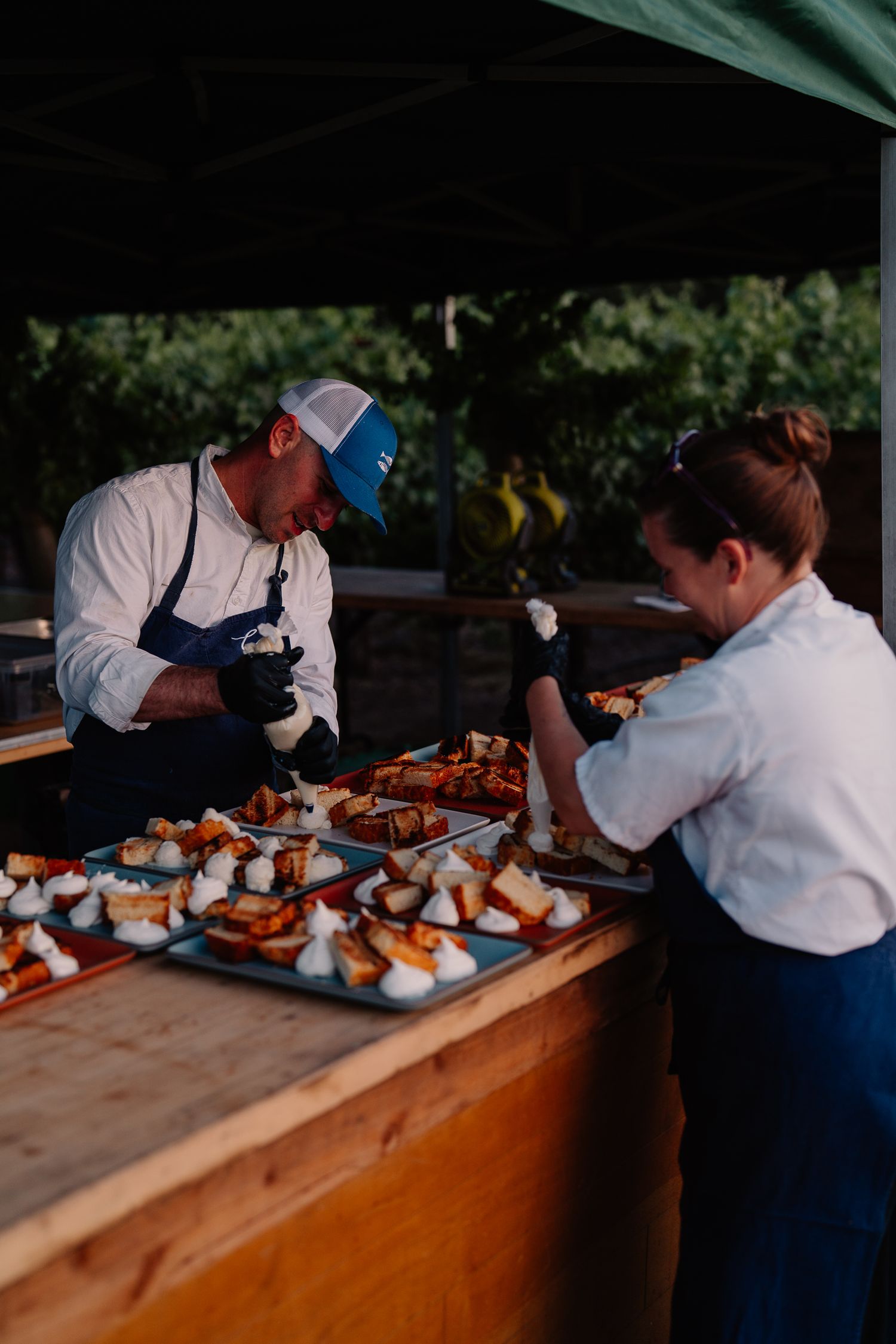 Chefs in white uniforms prepare appetizers on wooden boards at an outdoor catering station.