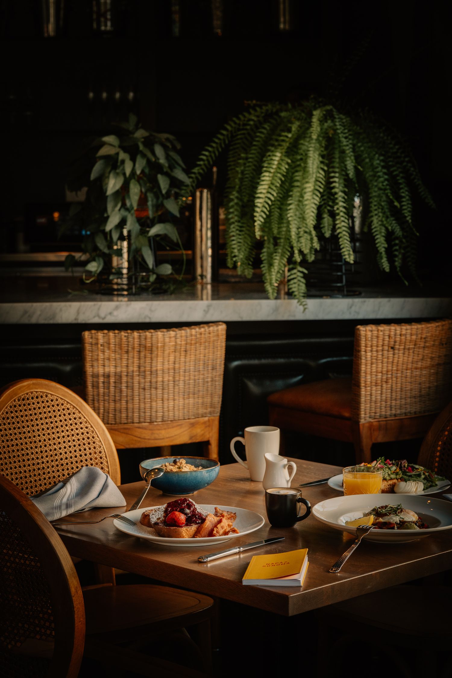 A cozy restaurant table setting with breakfast plates and coffee cups under hanging ferns in moody lighting.