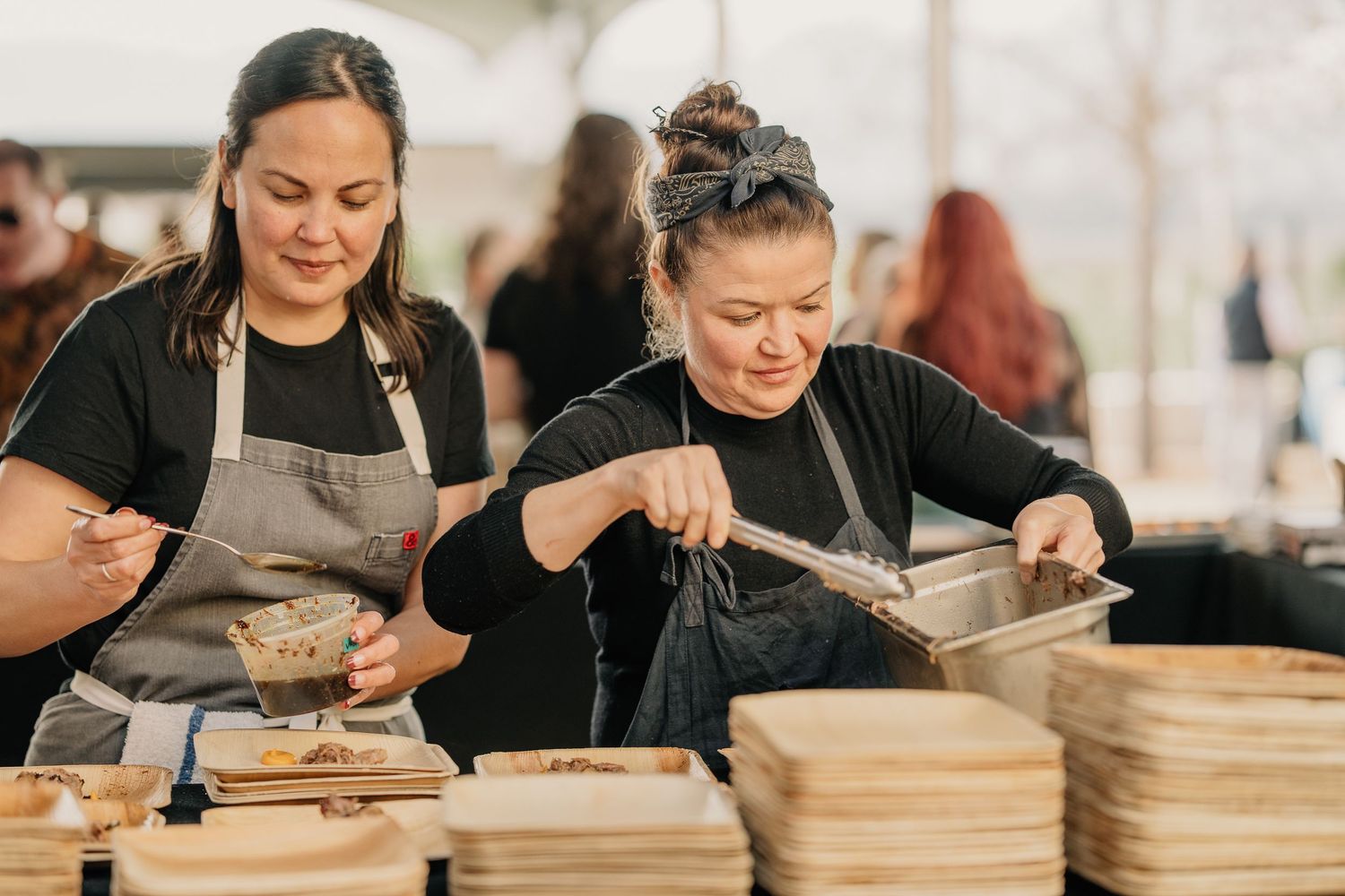 Two women in aprons serving food at an outdoor catering event.