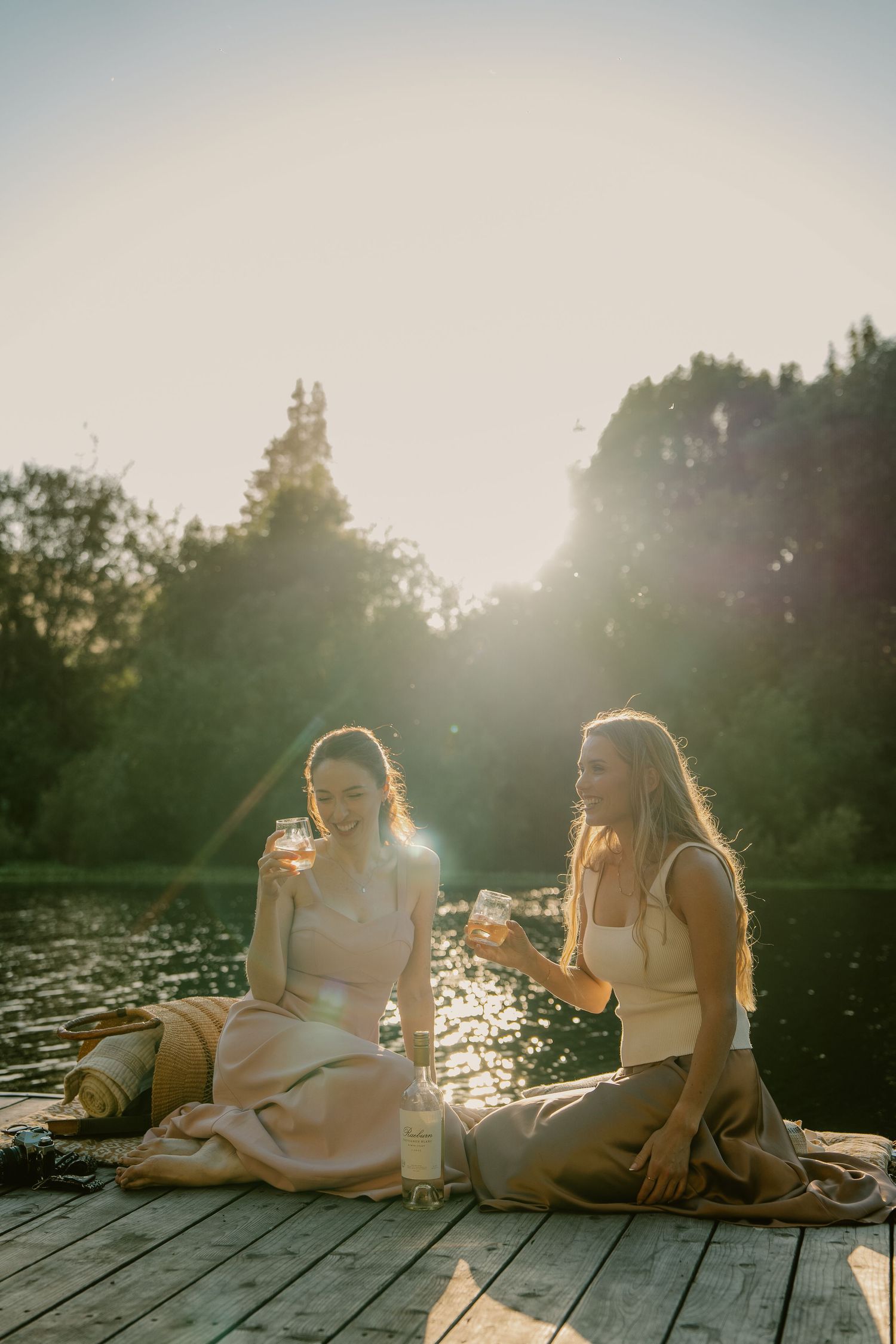 Two women sitting on a wooden dock by a lake, holding drinks and laughing in golden sunset light.