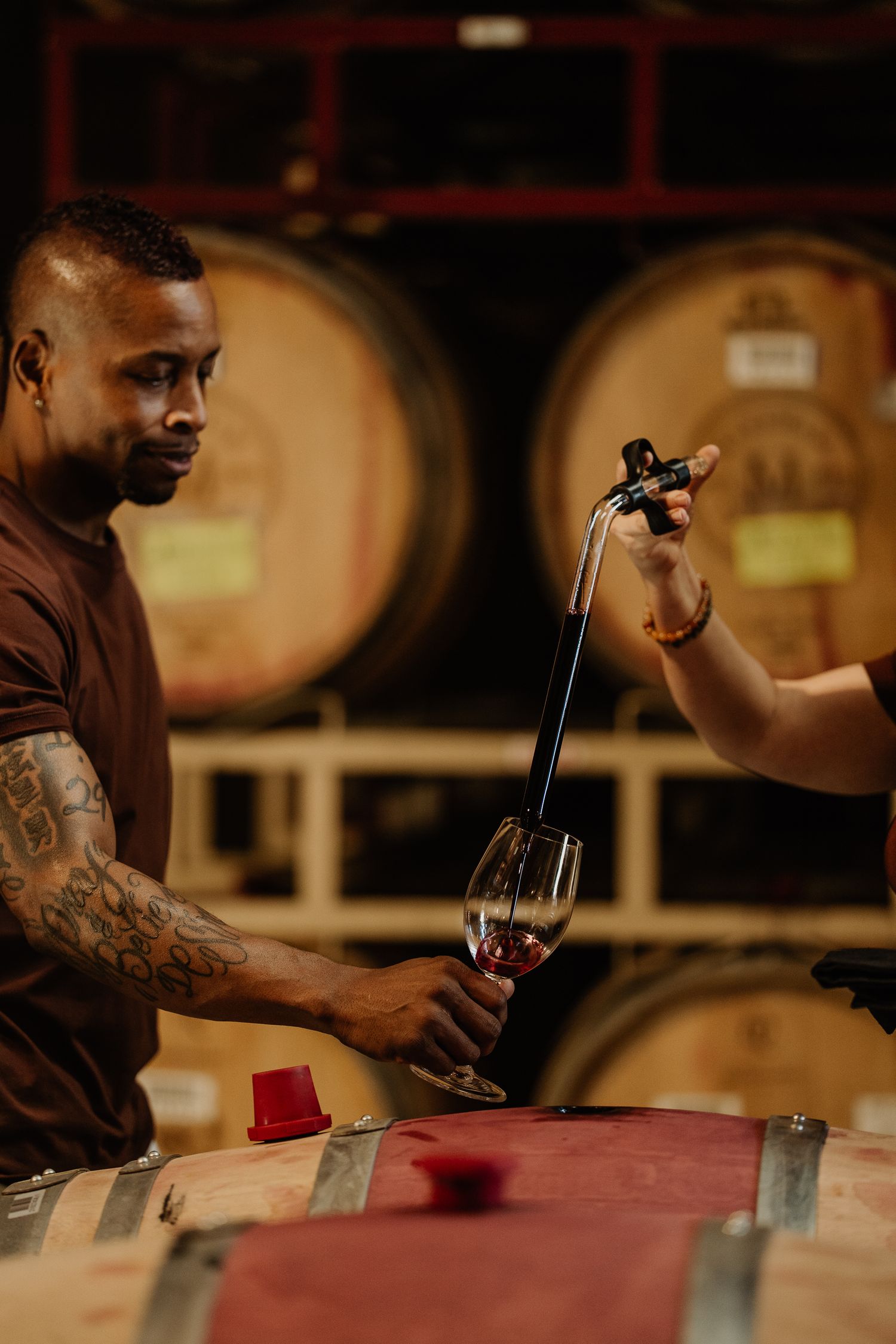 A tattooed man holds a wine glass while another person pours red wine using a wine thief in a barrel cellar.