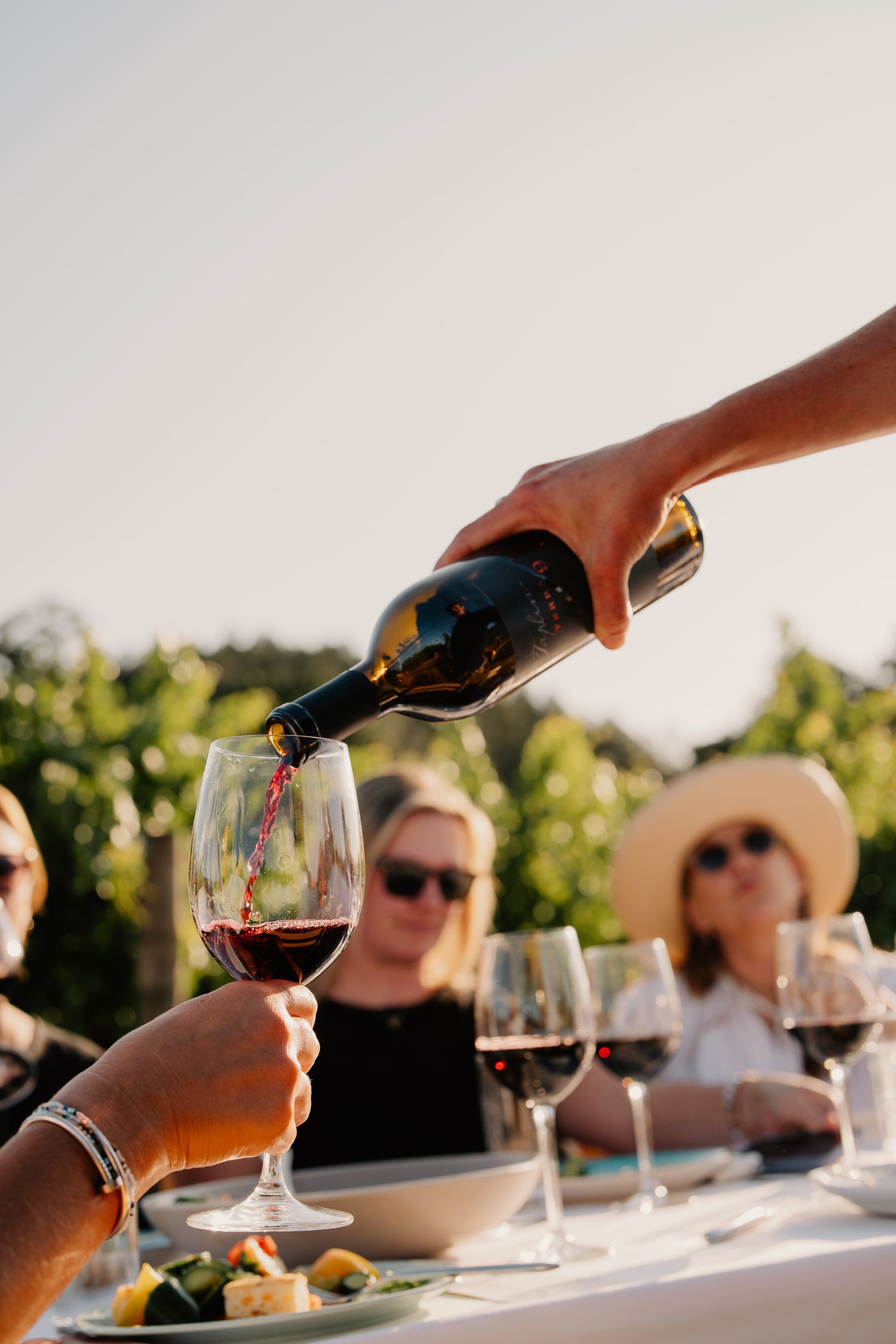 Red wine being poured into a glass at an outdoor vineyard tasting with guests seated at a table.