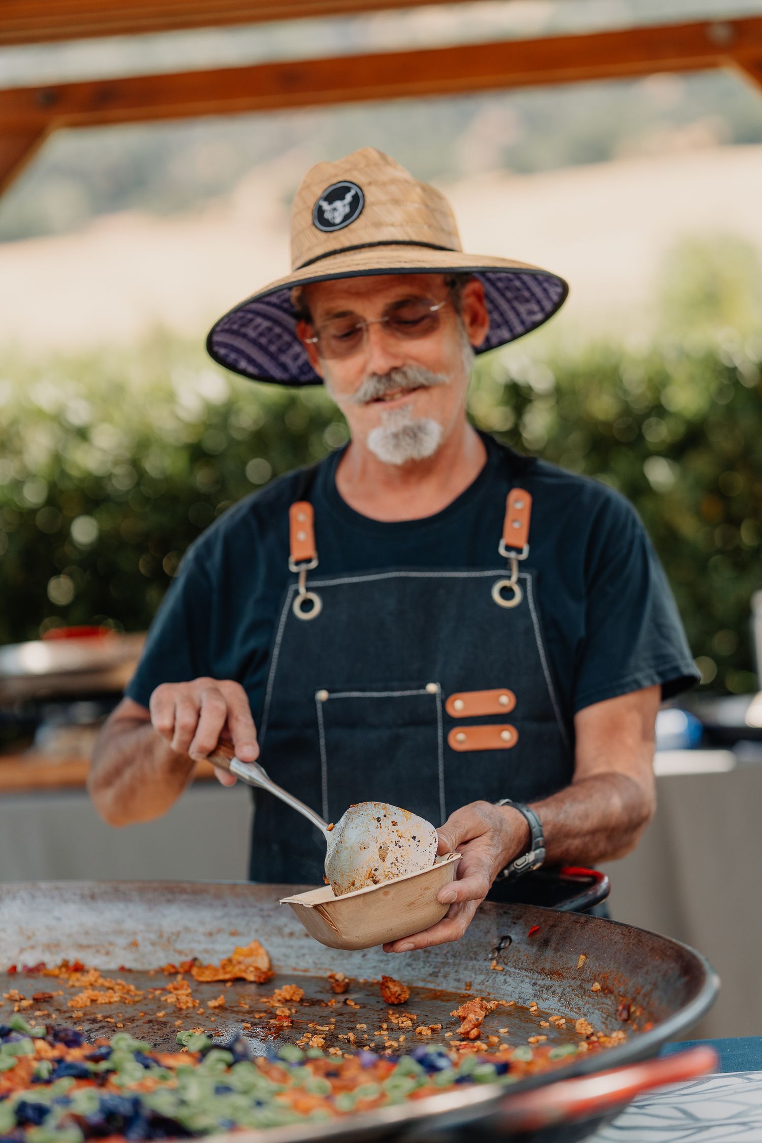 Chef in straw hat and apron serving paella from a large pan at an outdoor Silver Oak winery event.