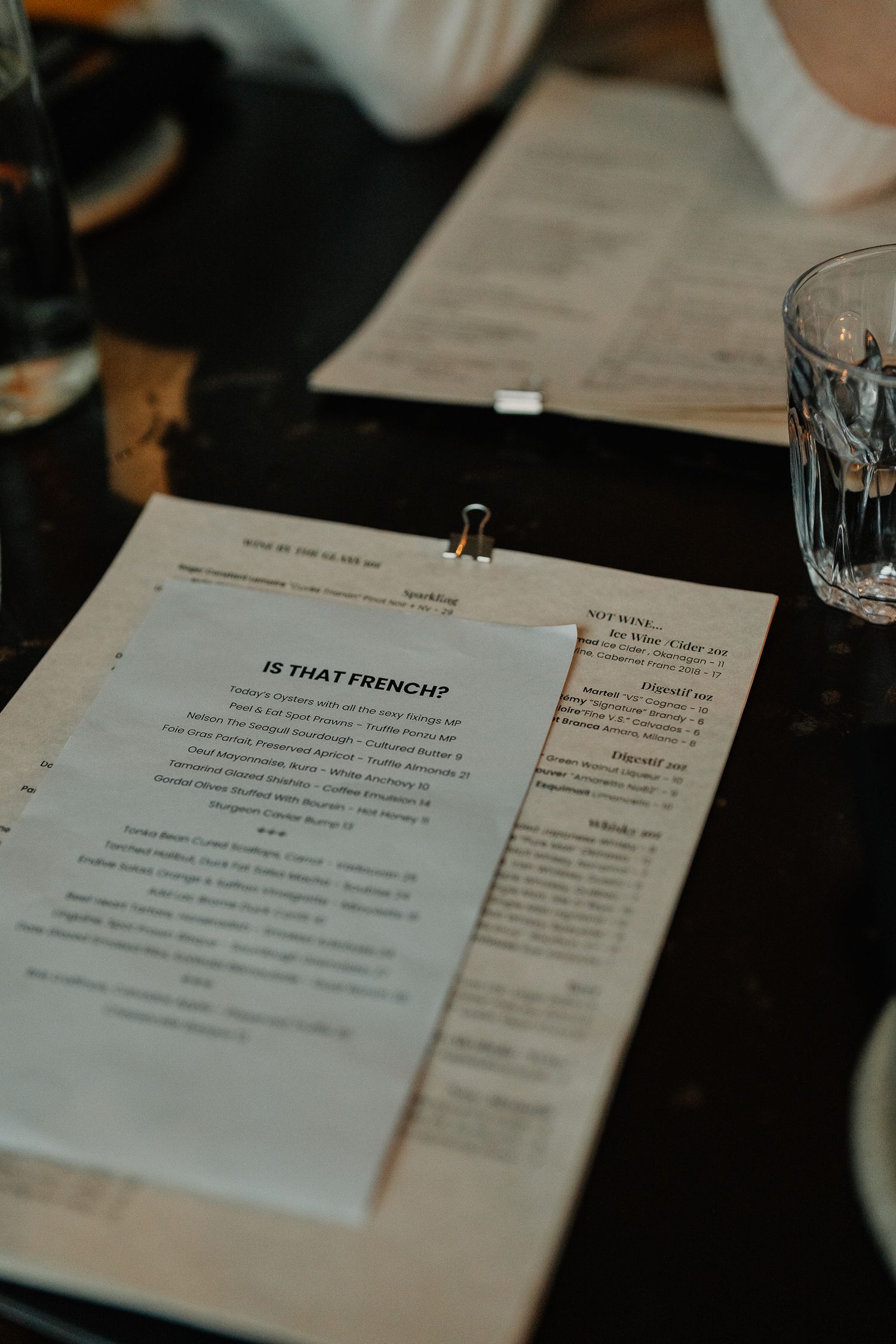 Restaurant menu titled 'IS THAT FRENCH?' on a clipboard with glass of water on dark wooden table in dim lighting.