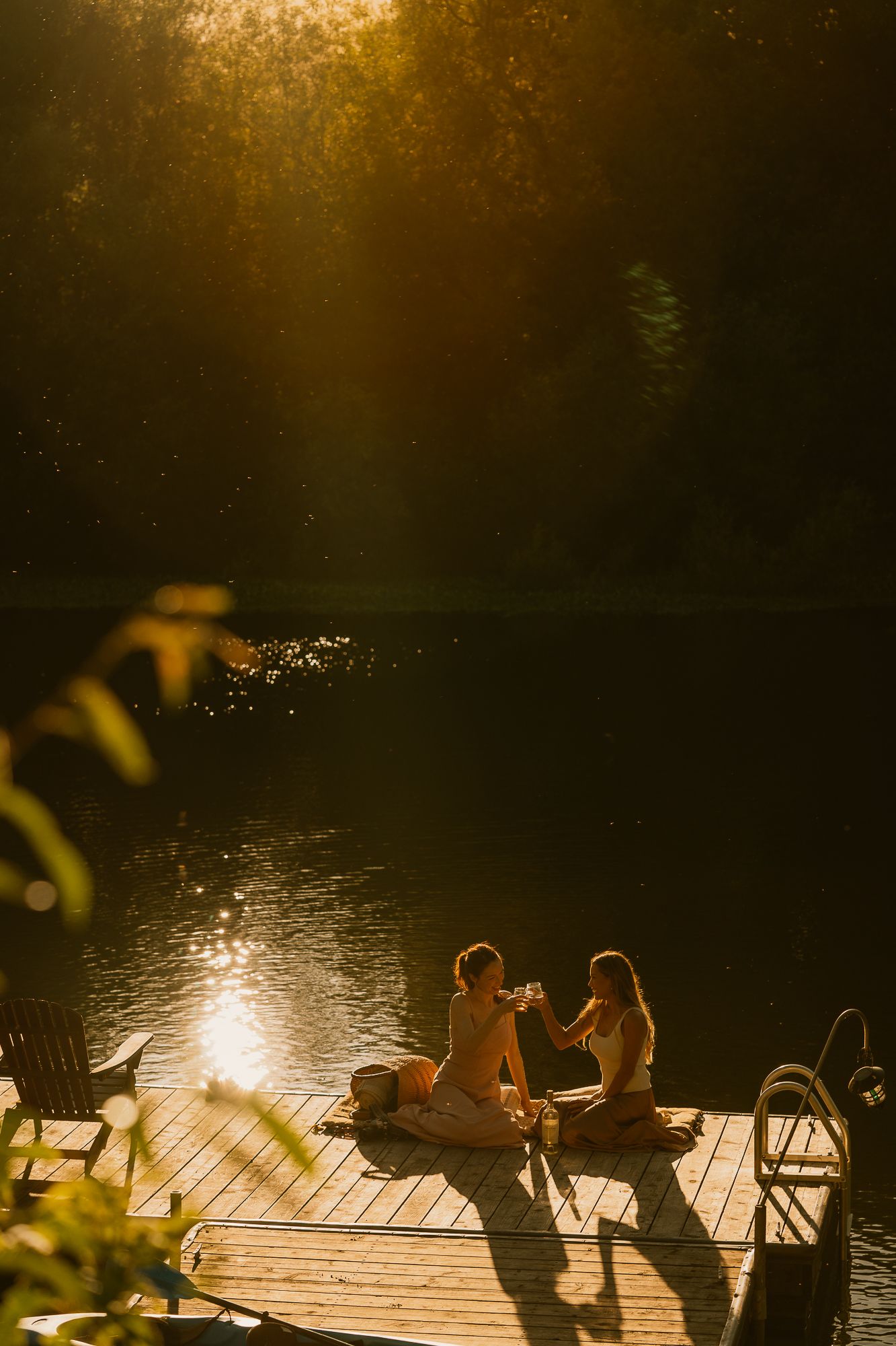 Two women sitting on a wooden dock by a lake, sharing drinks in golden sunset light surrounded by lush trees.