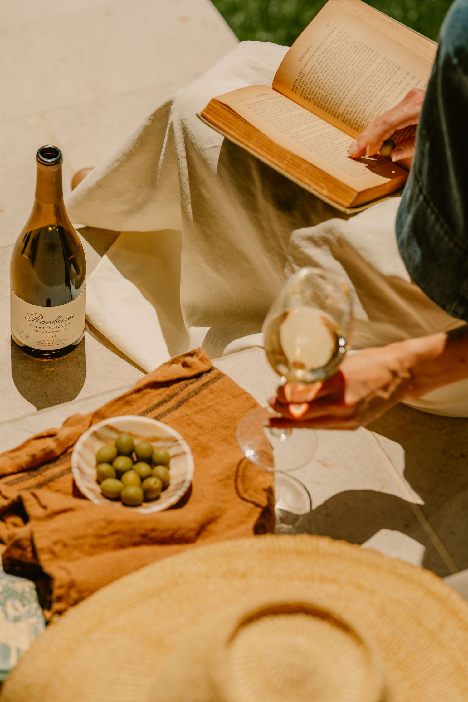 Outdoor picnic scene with white wine, olives, a straw hat, and an open book on a linen blanket.