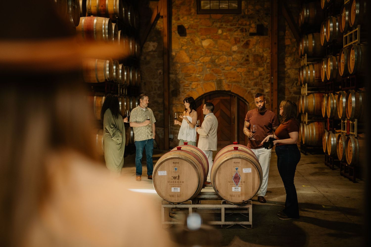 Group of people tasting wine in a rustic barrel cellar with stone walls and oak barrels.