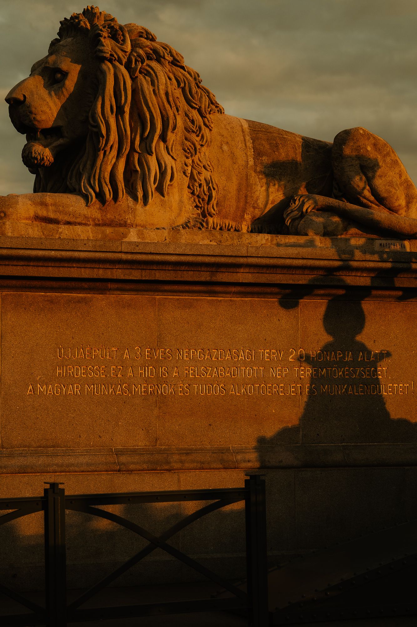 Stone lion sculpture on a pedestal with Hungarian inscription, bathed in golden sunset light.