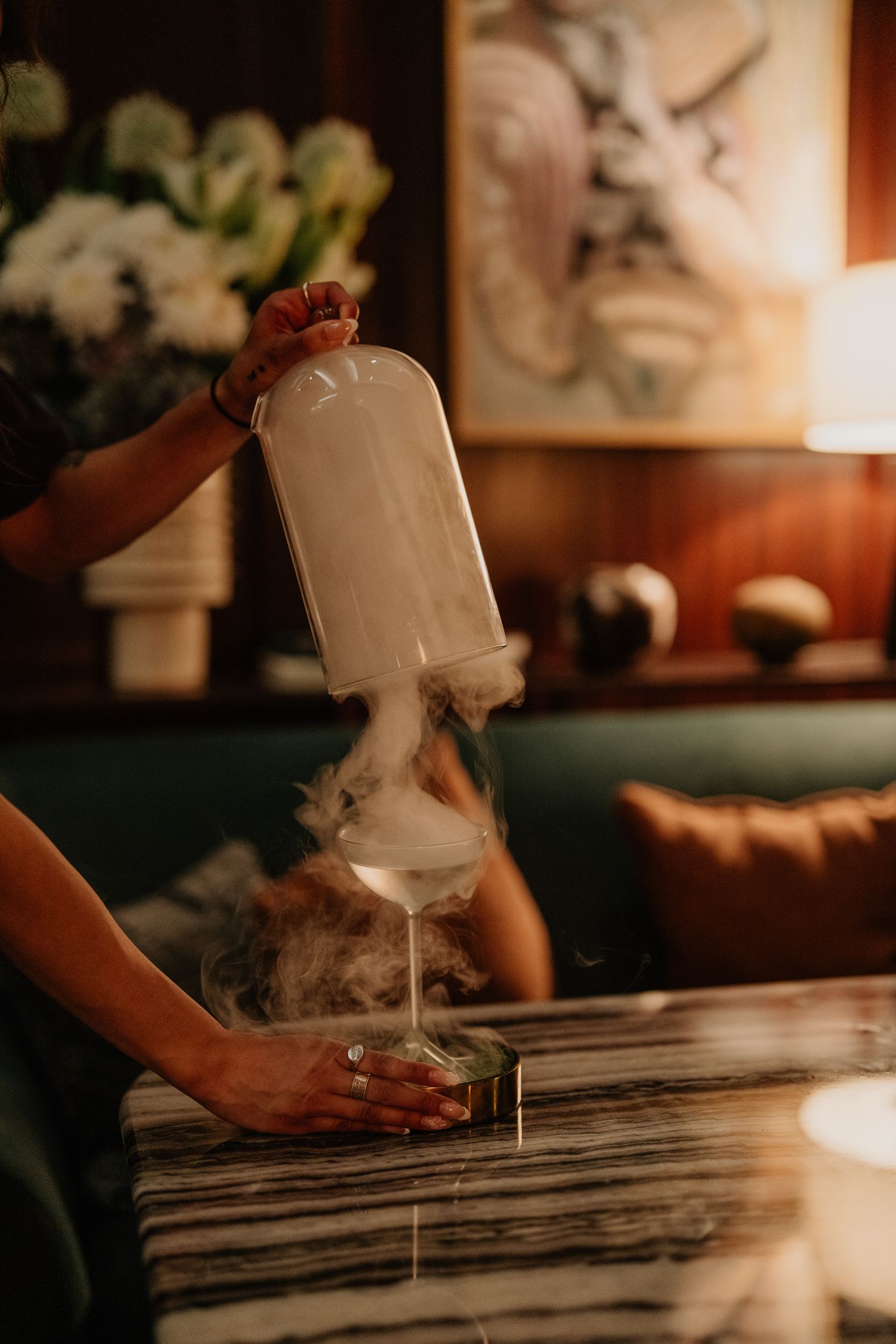 Hands pouring a creamy white liquid from a glass bottle onto a rustic wooden table surface in warm lighting.