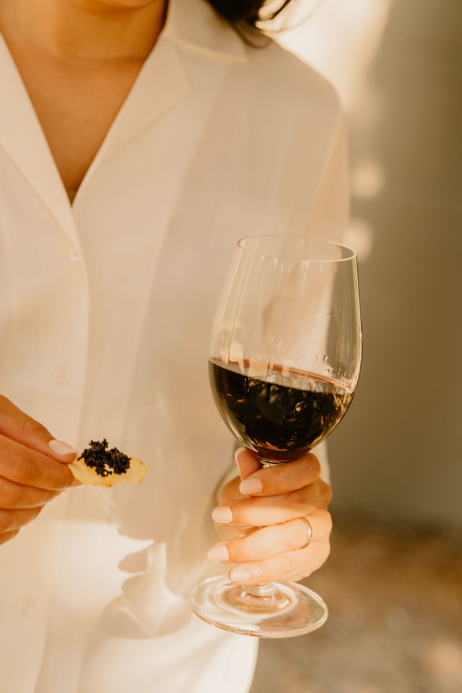 Woman in white shirt holding a glass of red wine and a caviar appetizer in warm sunlight.