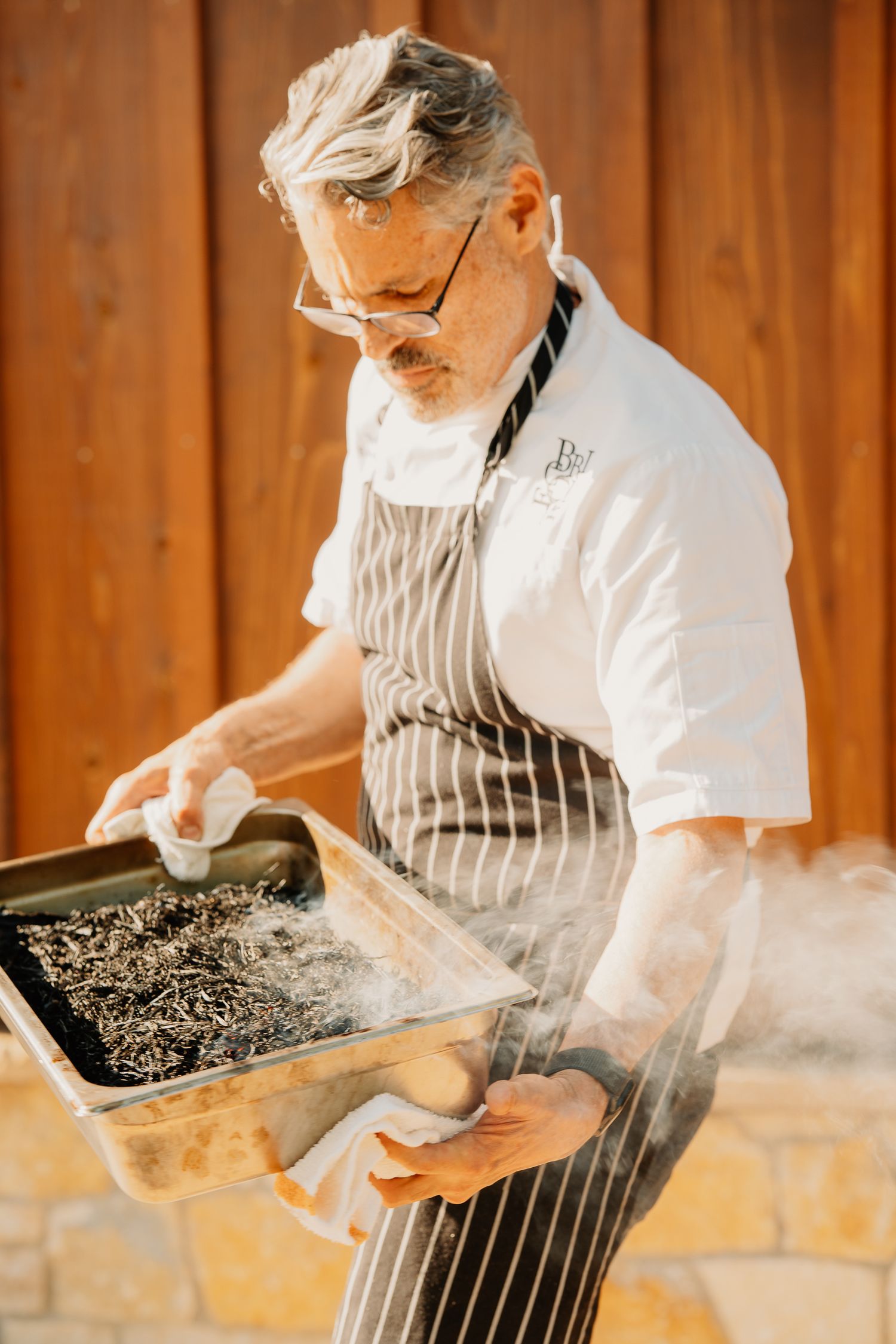 Chef in striped apron and white coat inspects steaming tray of food in rustic kitchen setting with wooden wall.