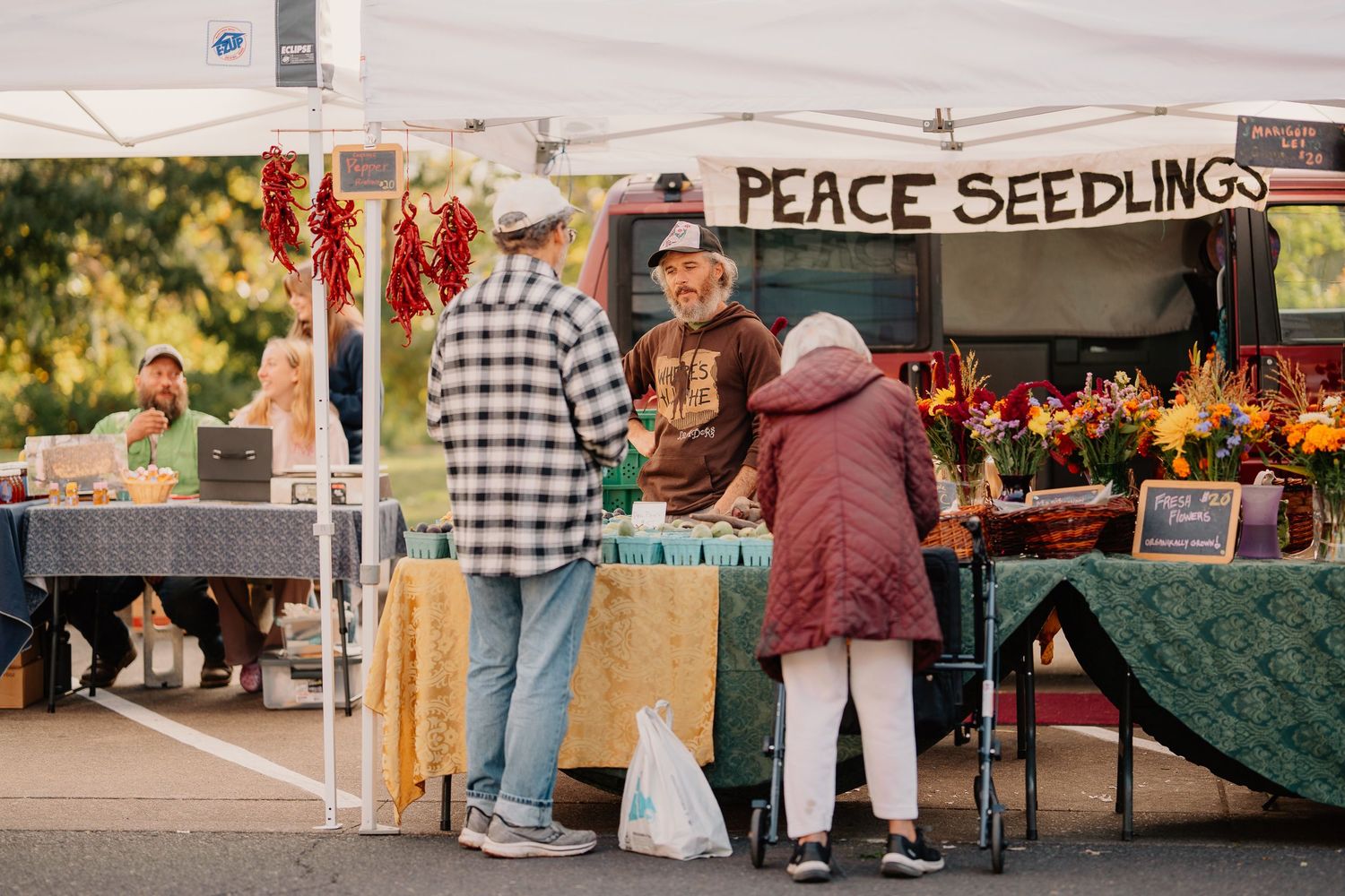 Customers browse a flower and seedling stall at an outdoor farmers market with colorful hanging plants on display.
