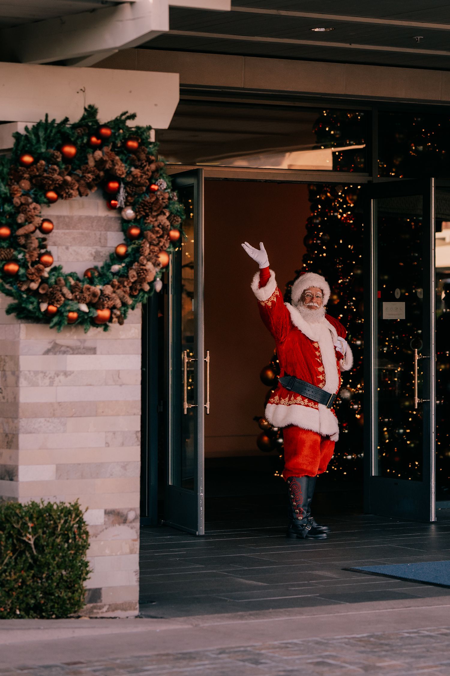 A Santa Claus figure waves while climbing up a wall outside a building decorated with a Christmas wreath.