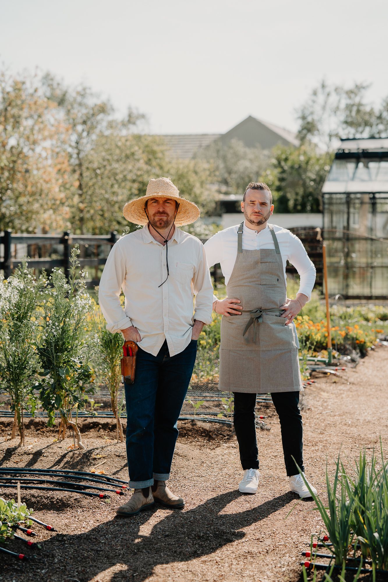 Two men stand in a lush garden, one wearing a straw hat and one in a chef's apron, surrounded by herbs.