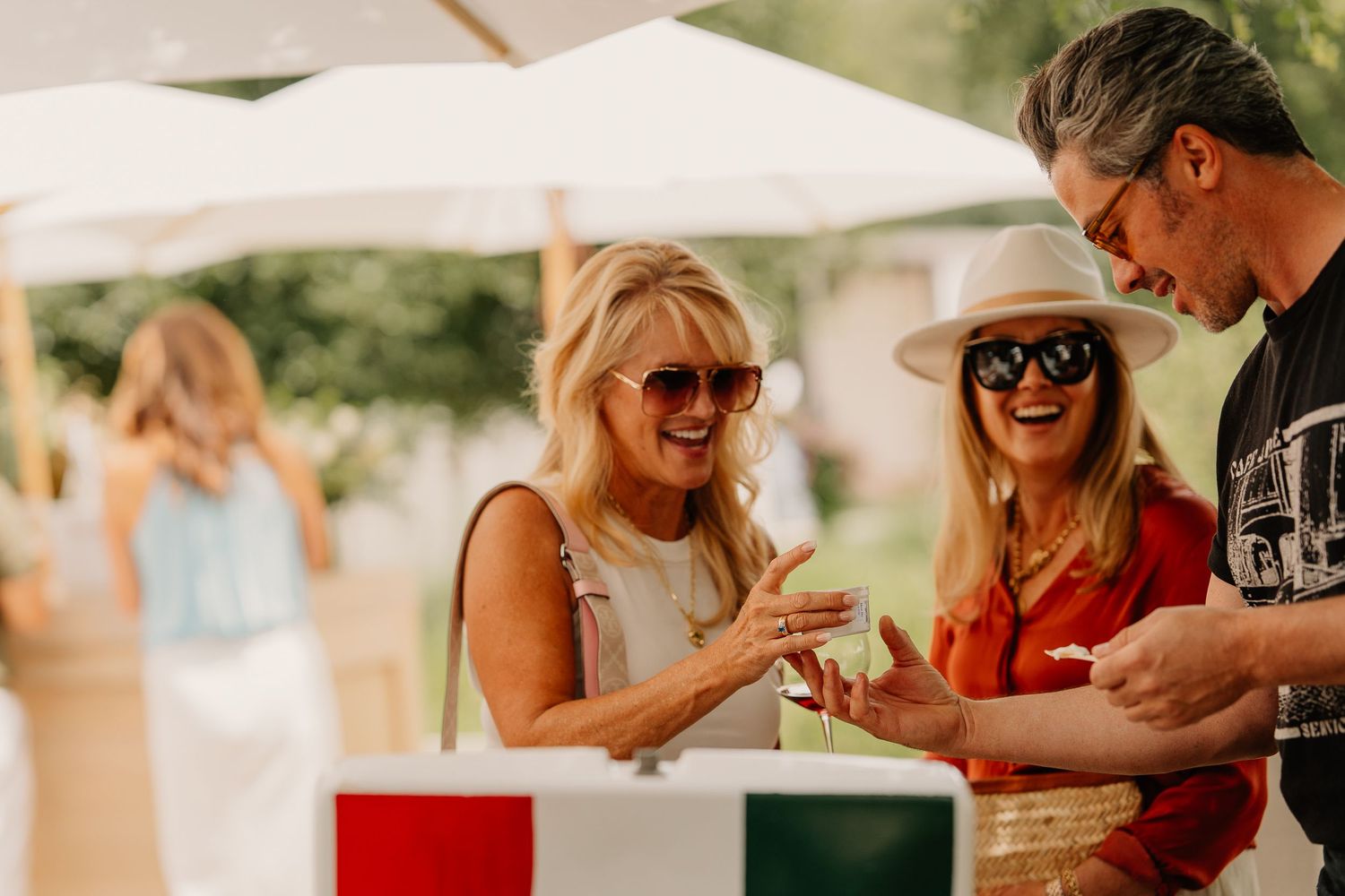 Group of friends enjoying drinks and conversation under white umbrellas at an outdoor summer event.
