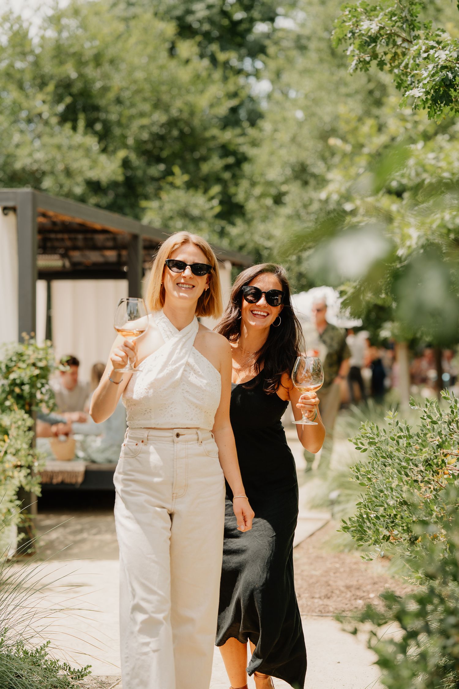 Two friends enjoy wine and conversation while strolling through a lush garden path on a sunny afternoon.