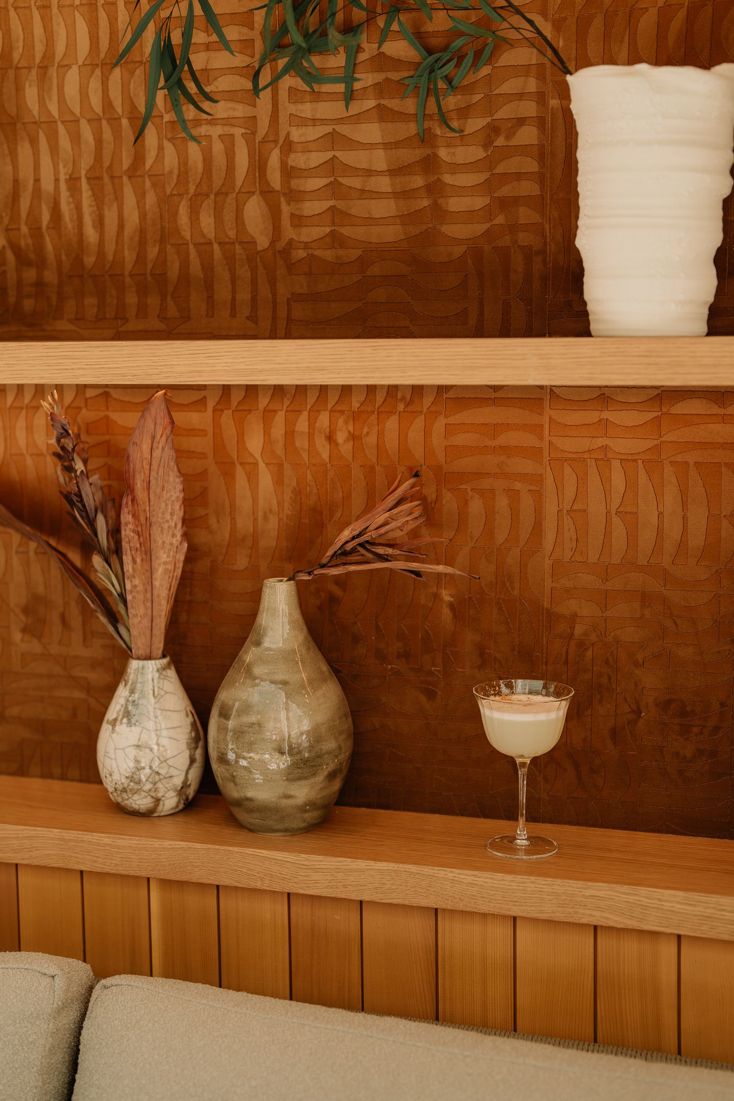 Wooden shelves displaying ceramic vases with dried flowers and a cocktail glass against a textured brown wall.