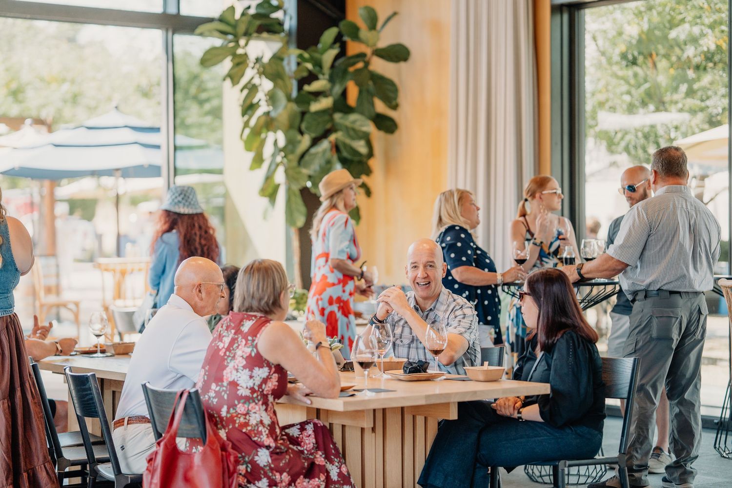 Guests dining and socializing at a bright, modern restaurant with large windows and lush greenery.
