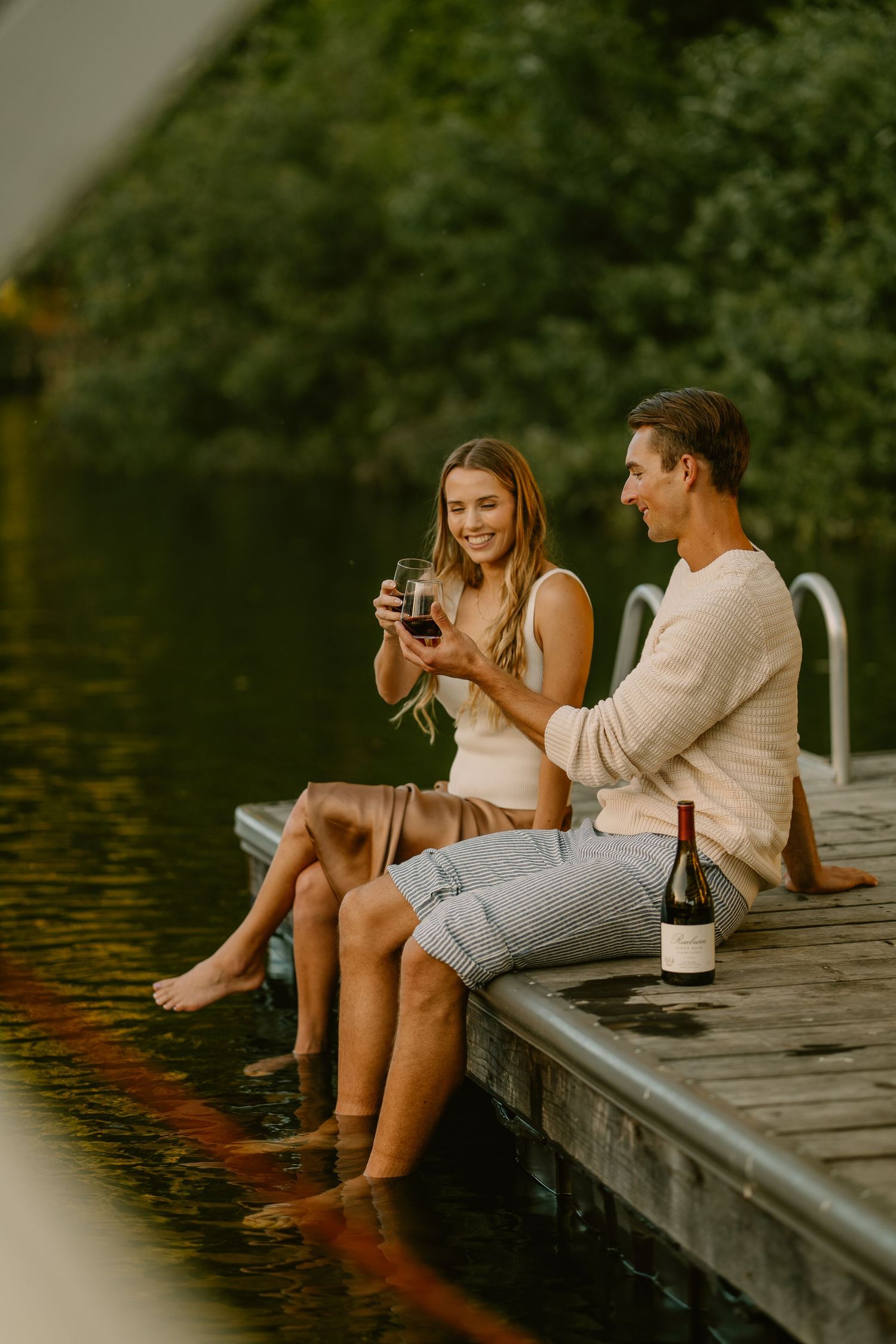 Couple sitting on a wooden dock sharing wine glasses with a bottle of red wine beside them near a serene lake.