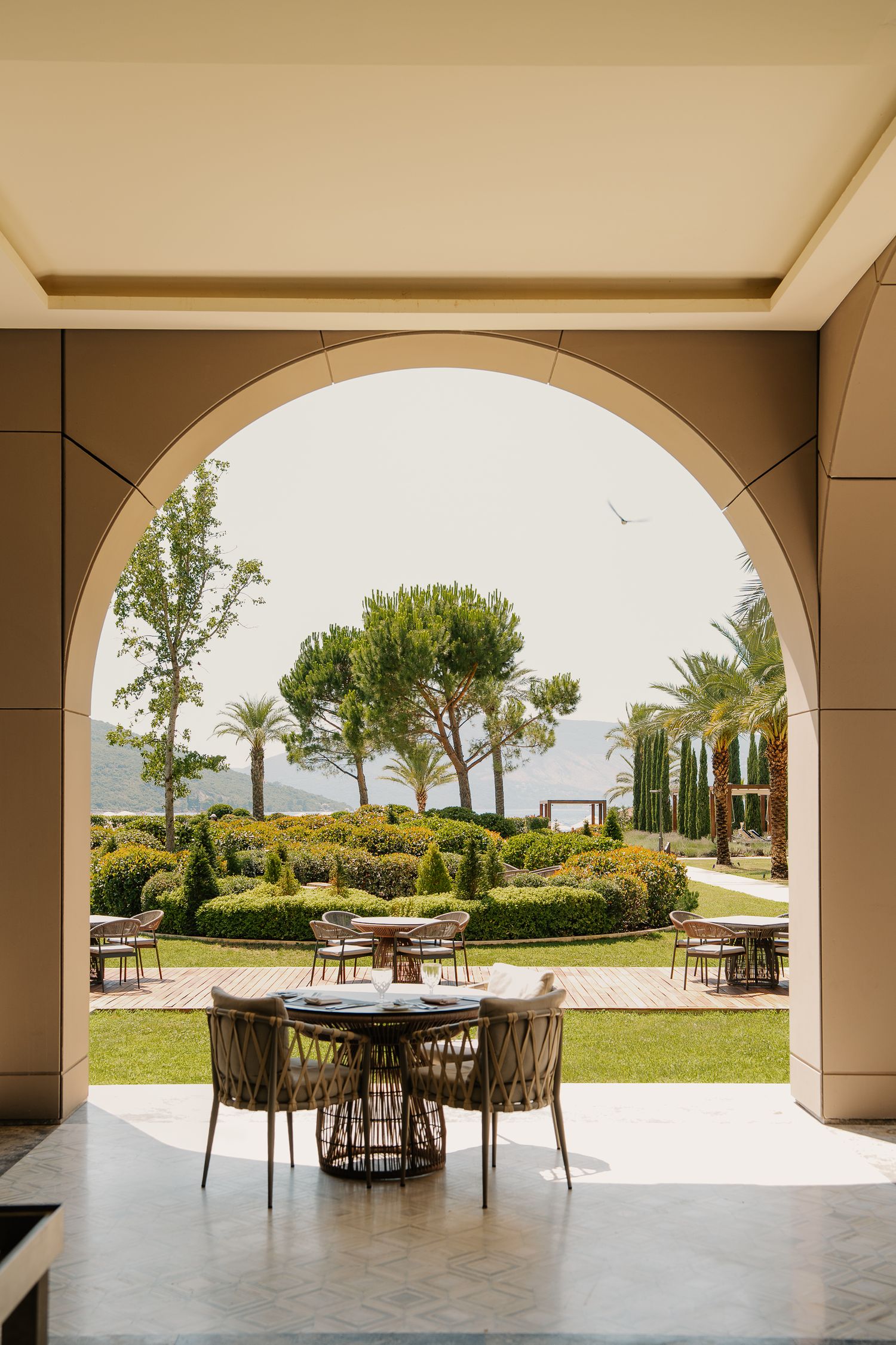 Elegant arched terrace overlooking a manicured garden with palm trees, outdoor dining tables, and a serene waterfront view.