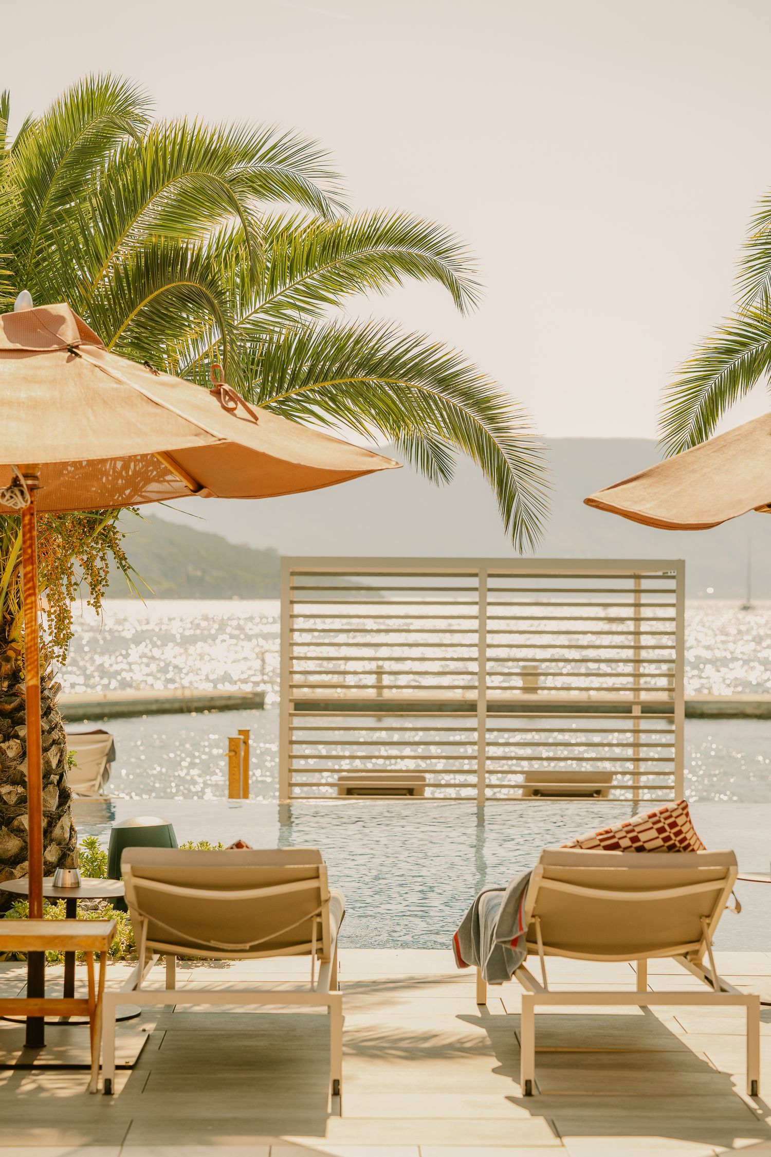 Luxury poolside lounge chairs with umbrellas and palm trees overlooking a sparkling Mediterranean bay.