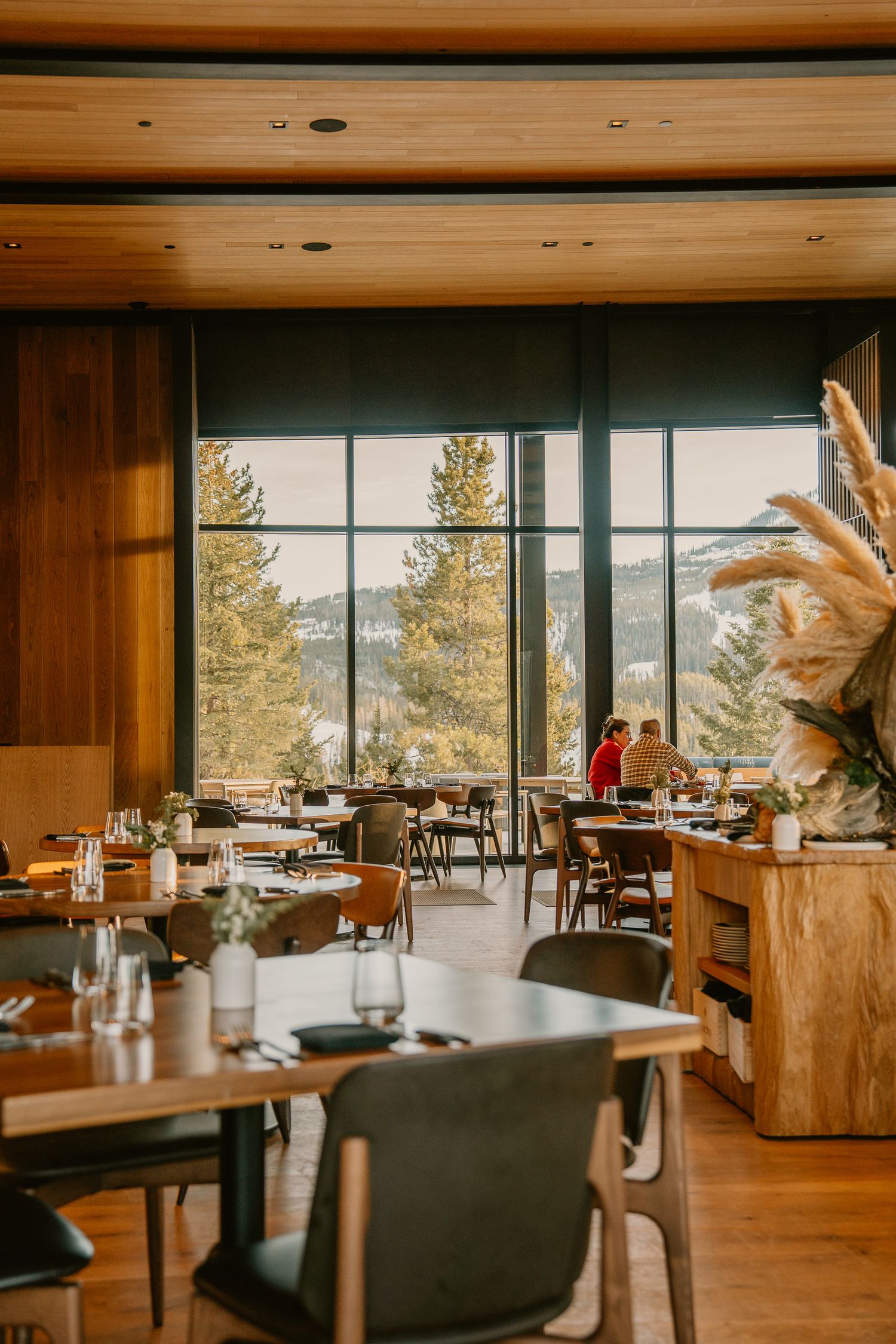 Modern restaurant interior with wooden walls and large windows offering panoramic mountain views and elegant table settings.